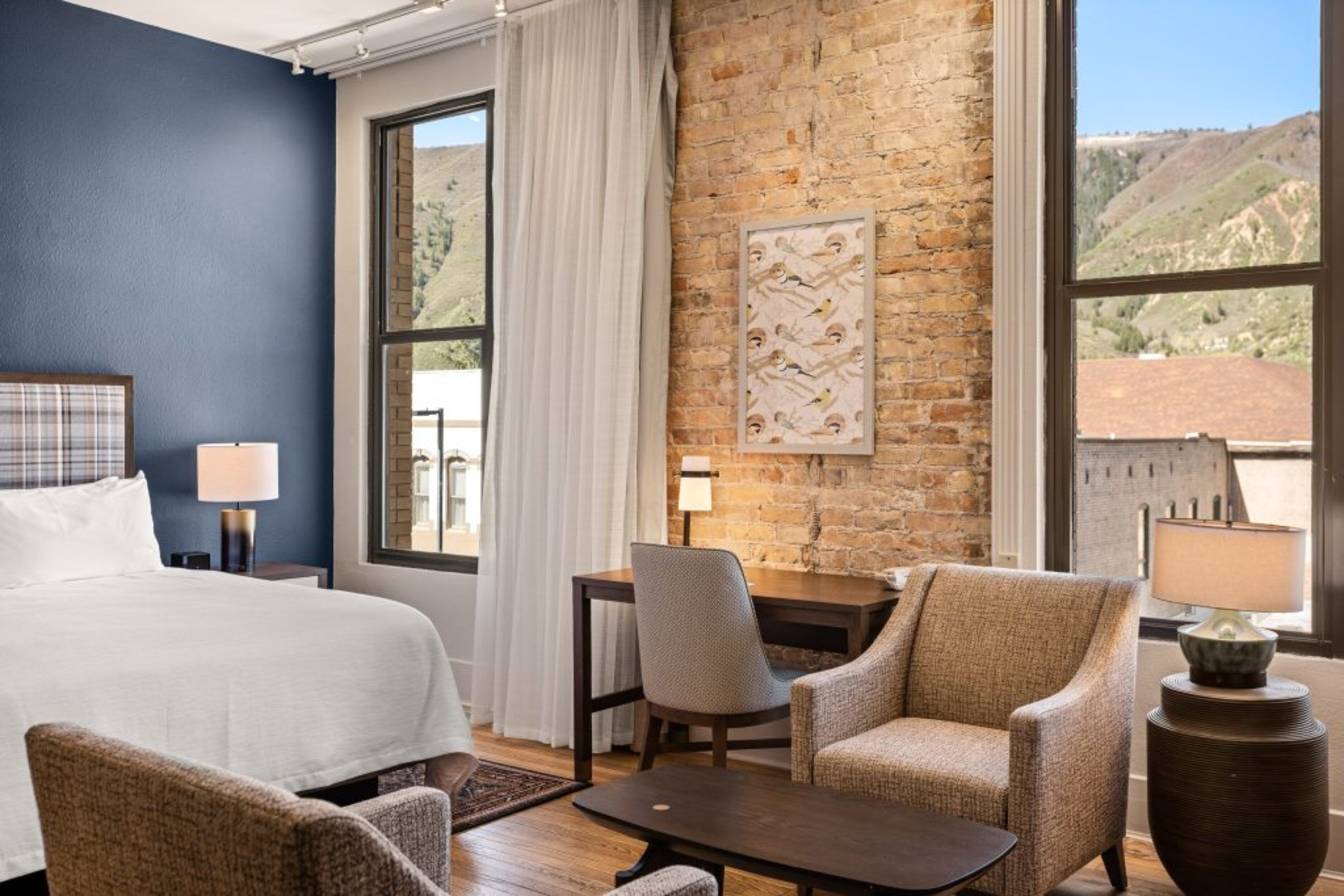 A hotel room with a large bed, two armchairs, a desk, and a view of a mountainous landscape through large windows.
