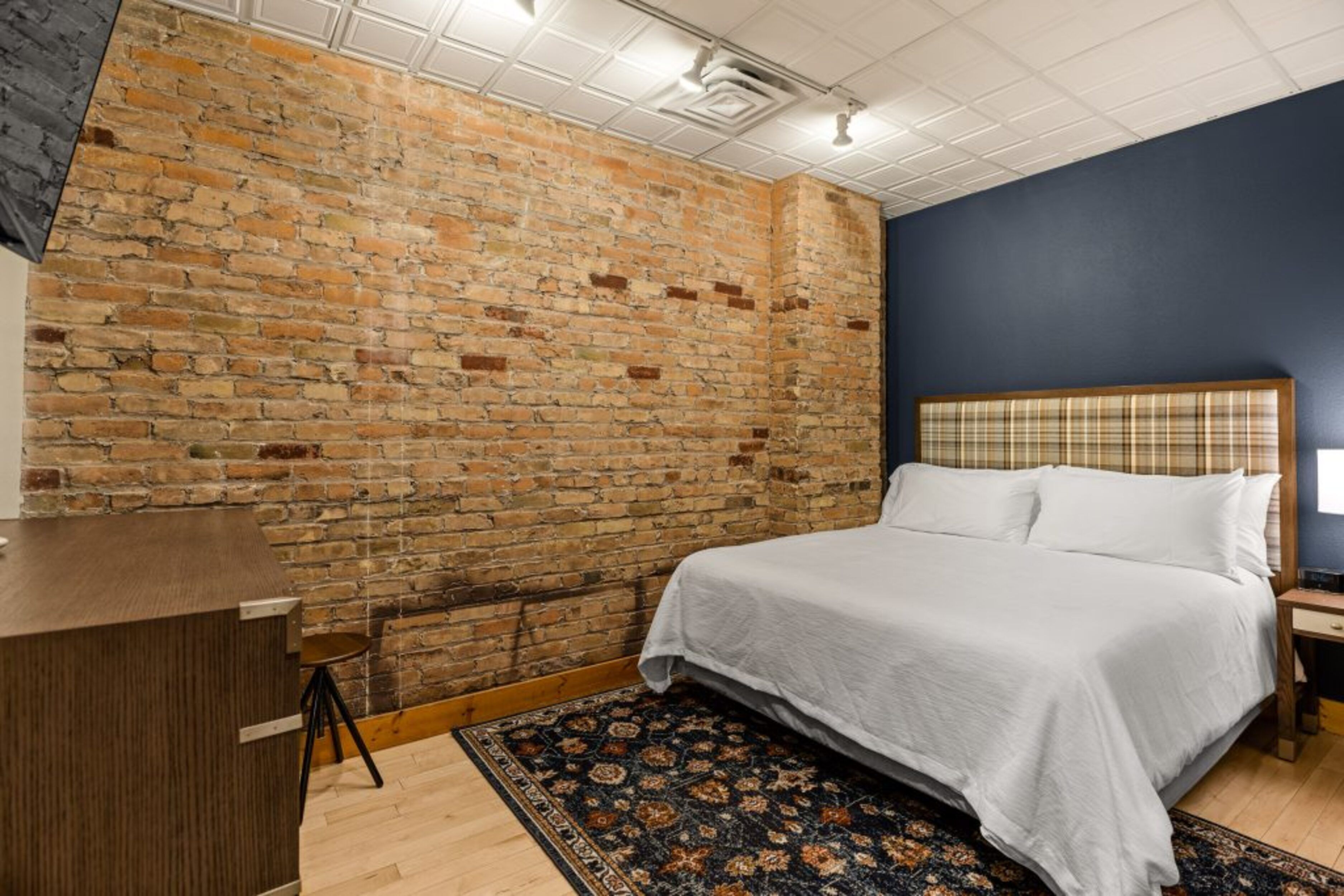 A bedroom with a brick wall, a bed with white linens, a wooden desk, and a patterned rug.