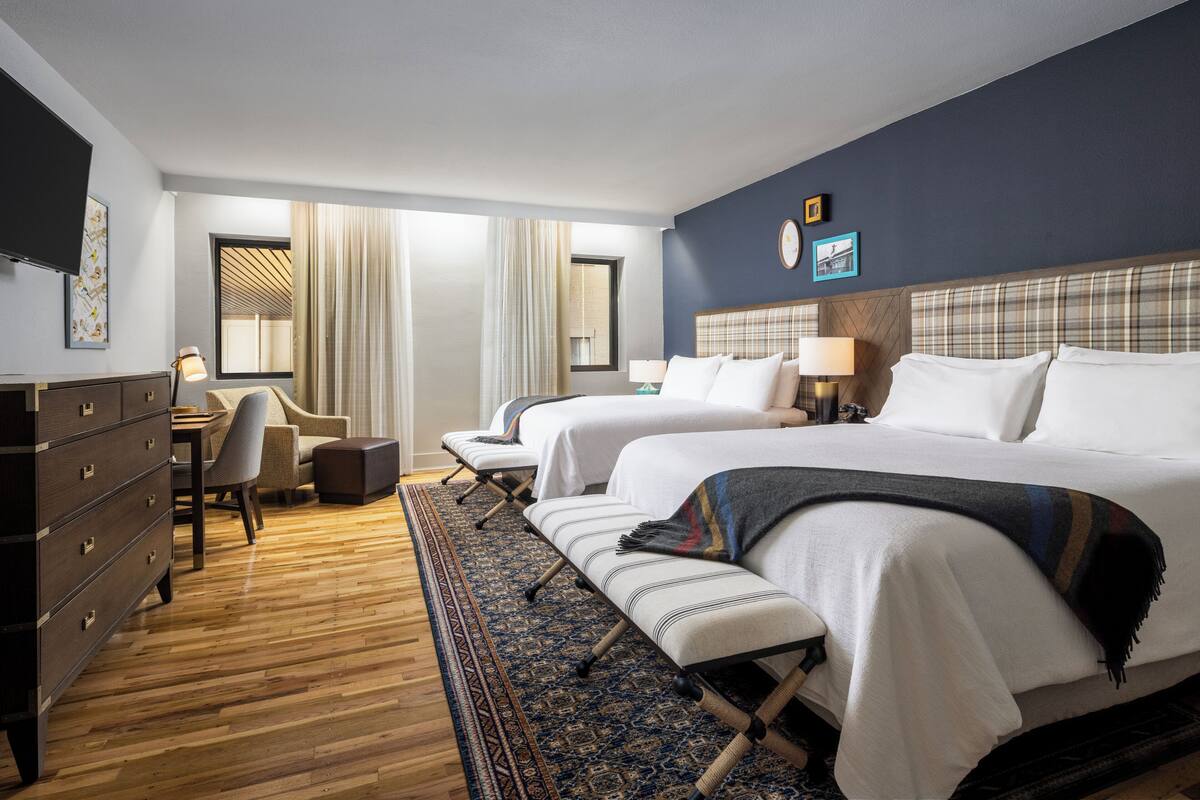 Habitación de hotel con dos camas, un escritorio, una silla, un aparador y un ventanal con cortinas.