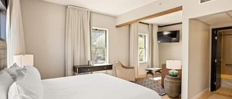 A bedroom with a large bed, a desk, a chair, a TV, and a door leading to another room.
