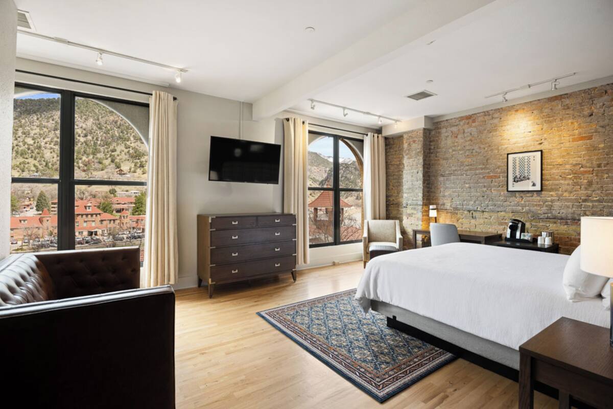 Una habitación de hotel moderna con una cama grande, un televisor de pantalla plana, un aparador y vistas a un paisaje montañoso a través de ventanas amplias.