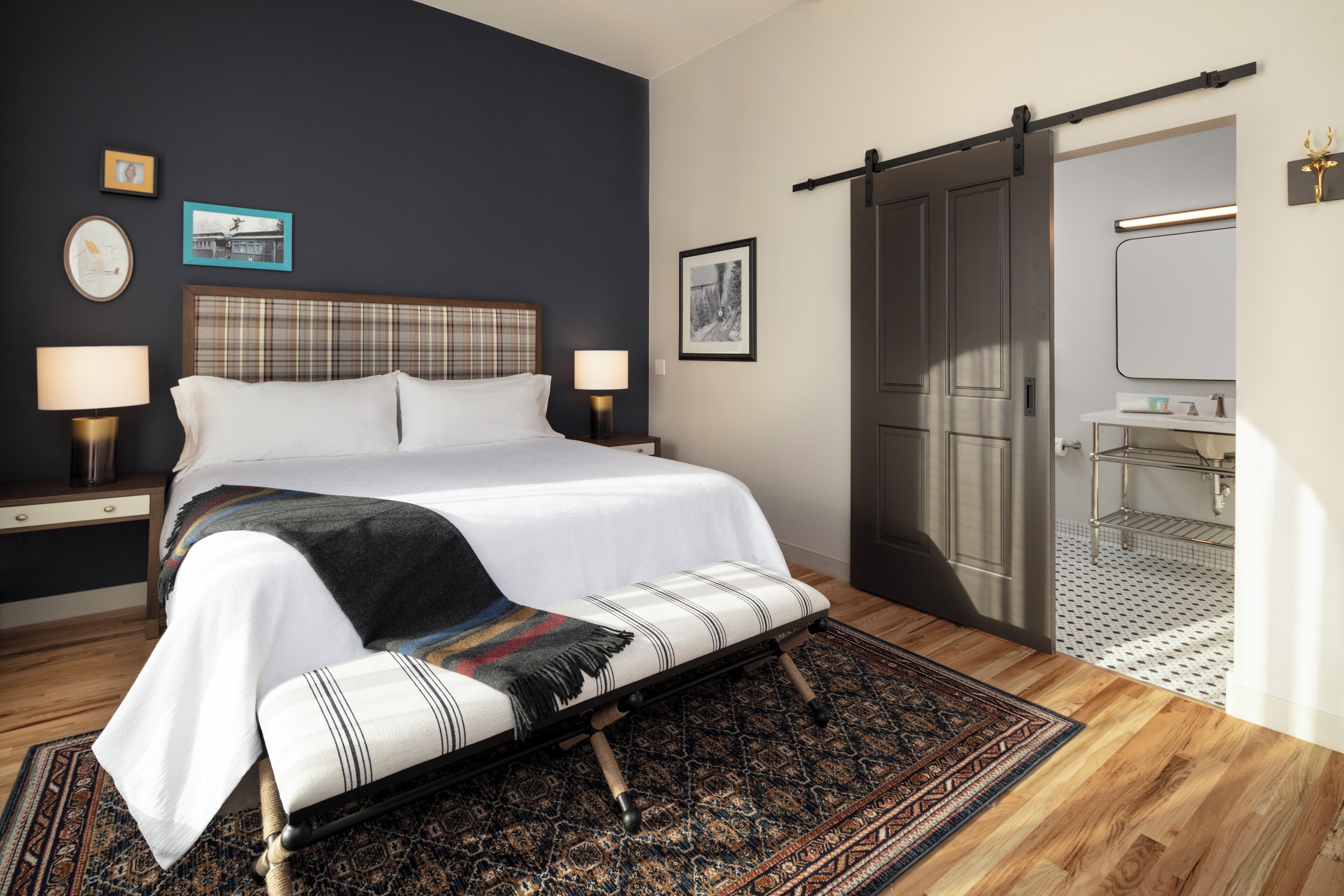 A bedroom with a bed, bedside lamps, a nightstand, a bench, a patterned rug, a dark wall, a door, and a small bathroom with a mirror and sink.