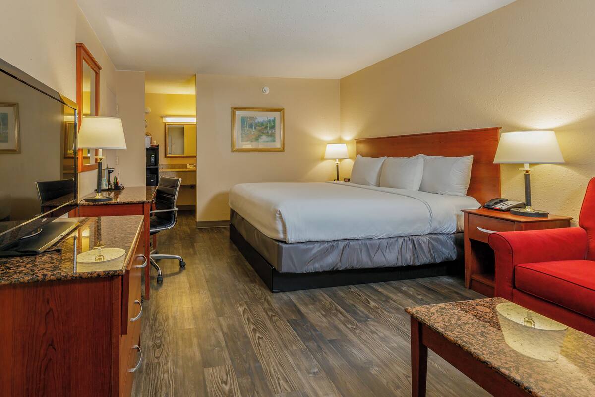 Une chambre d’hôtel avec un lit, un bureau avec un ordinateur, une chaise, un fauteuil rouge et une petite table.