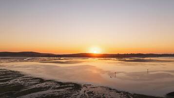 A wide view of a coastal landscape with a sunset over a calm sea, featuring a flat terrain with sparse vegetation and a distant horizon line.