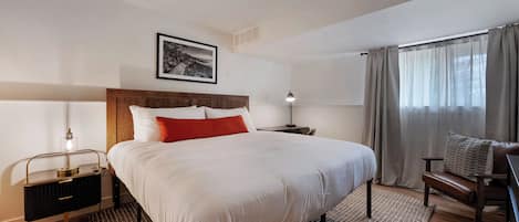 Quarto de hotel bem arrumado, com uma cama grande, uma mesa de cabeceira com abajur, uma cadeira e uma janela com cortinas.