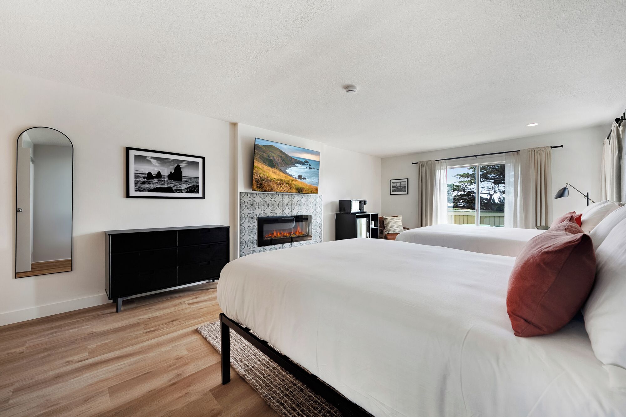 A modern hotel room with a fireplace, a bed with white bedding and a red pillow, a desk with a lamp, and a view of the outdoors through a window.