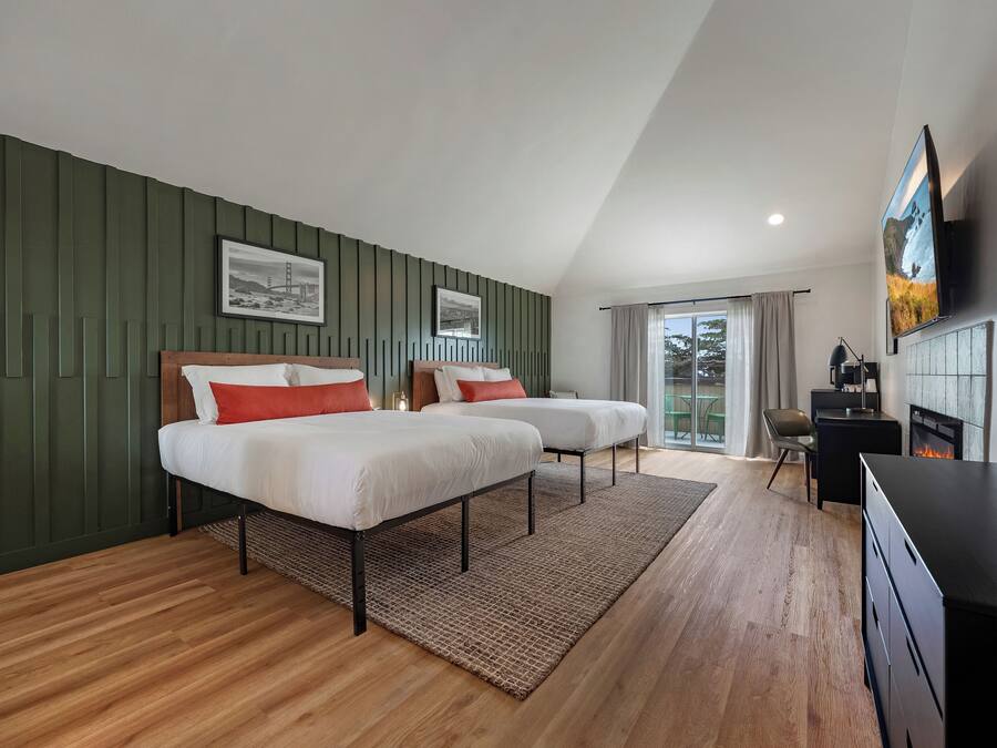A modern hotel room with two beds, a fireplace, a TV, and a view of the outdoors.