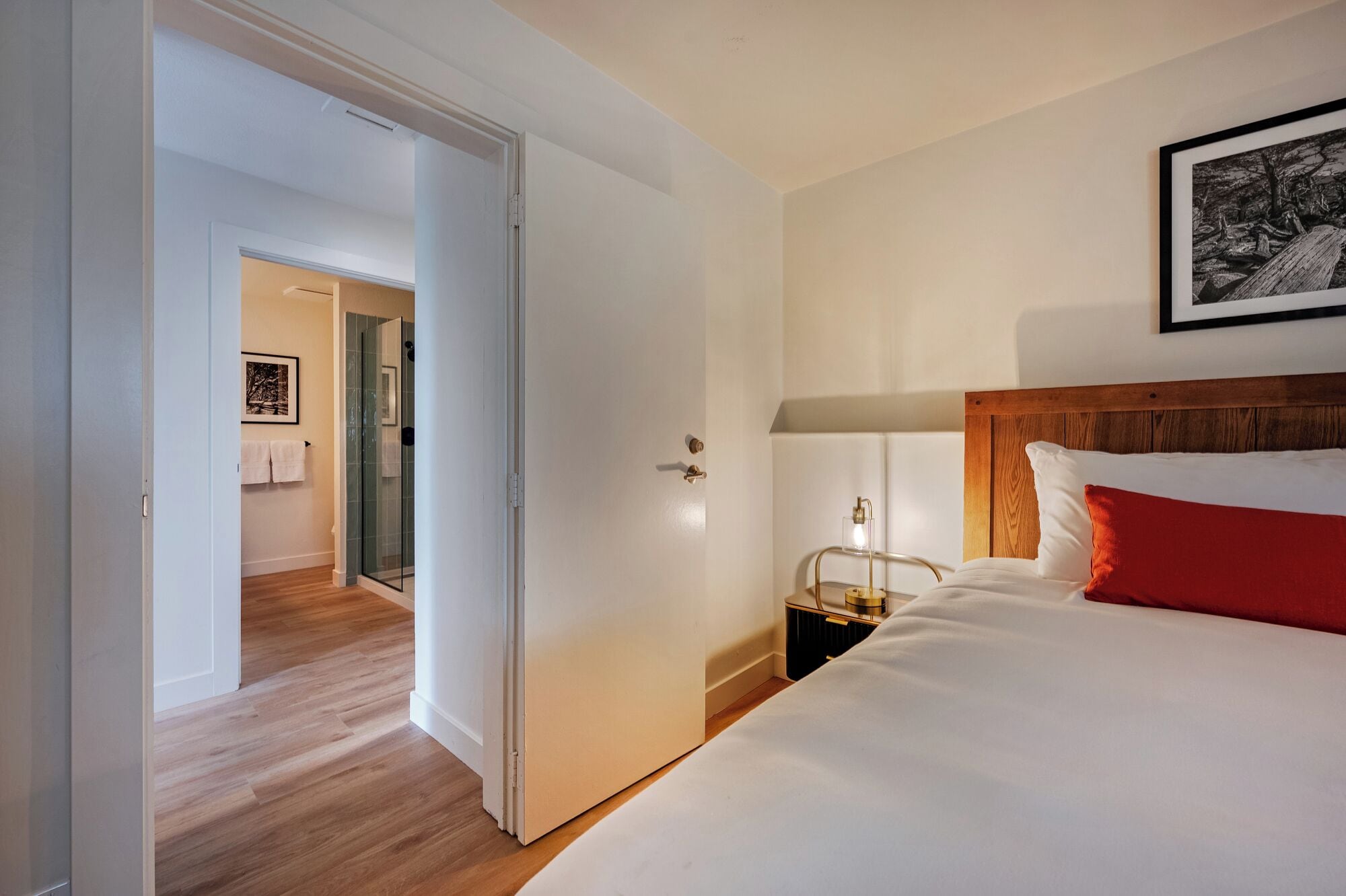 Ein Hotelzimmer mit Holzboden, einem Bett mit weißer und roter Bettwäsche, einem Nachttisch mit Lampe und einem gerahmten Bild an der Wand.