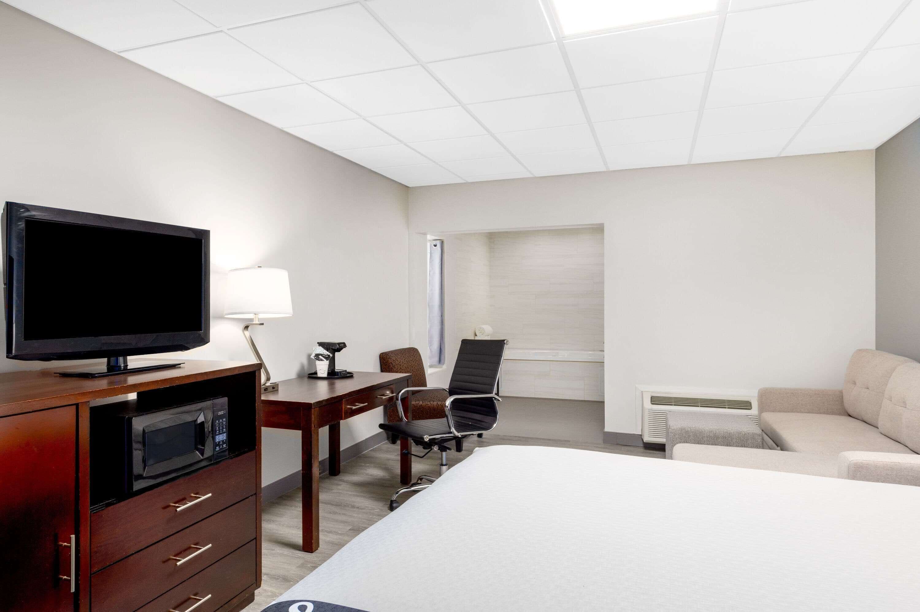 Quarto de hotel com cama, escrivaninha com computador, cadeira, televisão, luminária e ar condicionado.
