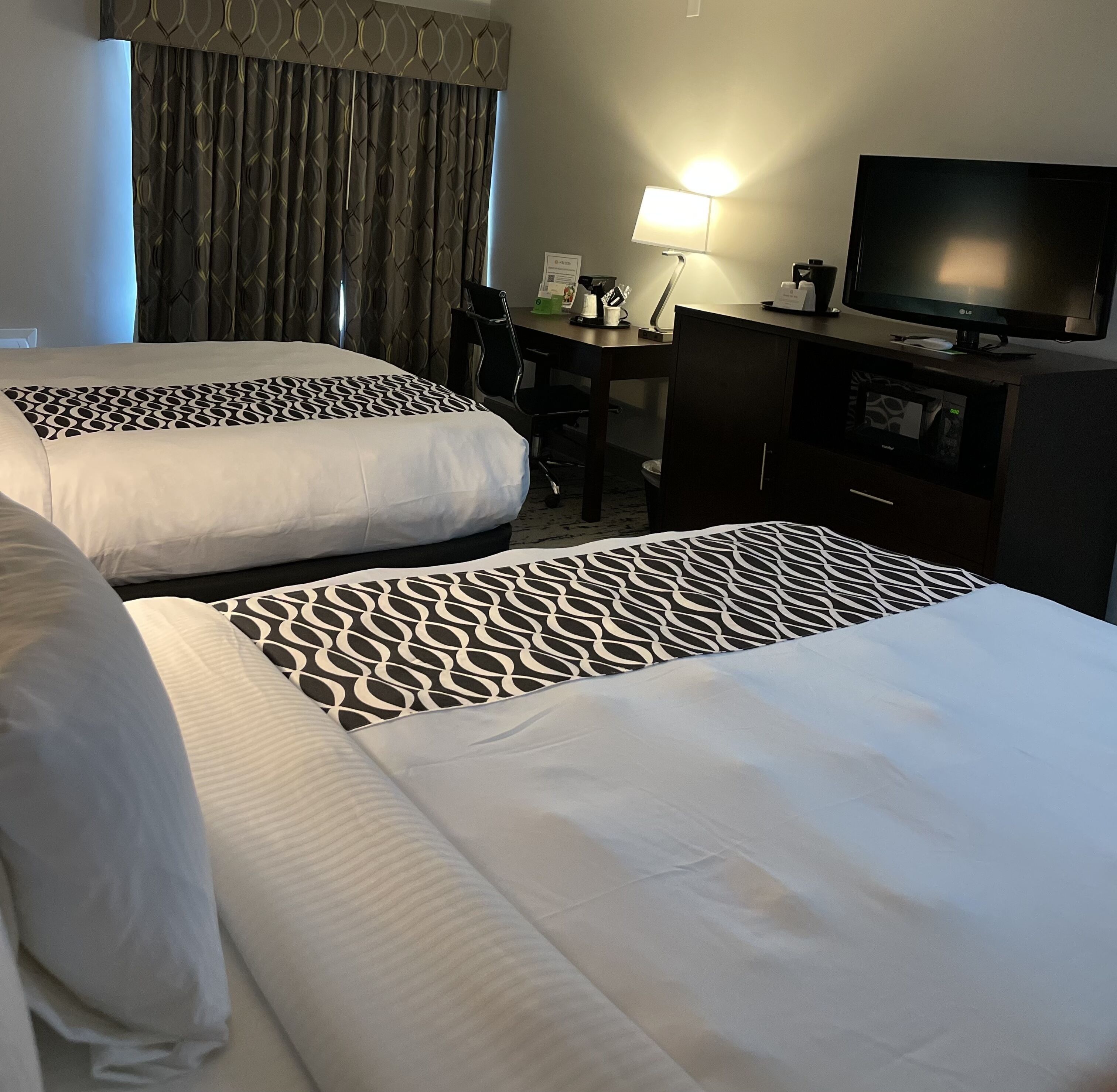 A hotel room with two beds, a desk, a chair, a TV, and a lamp.