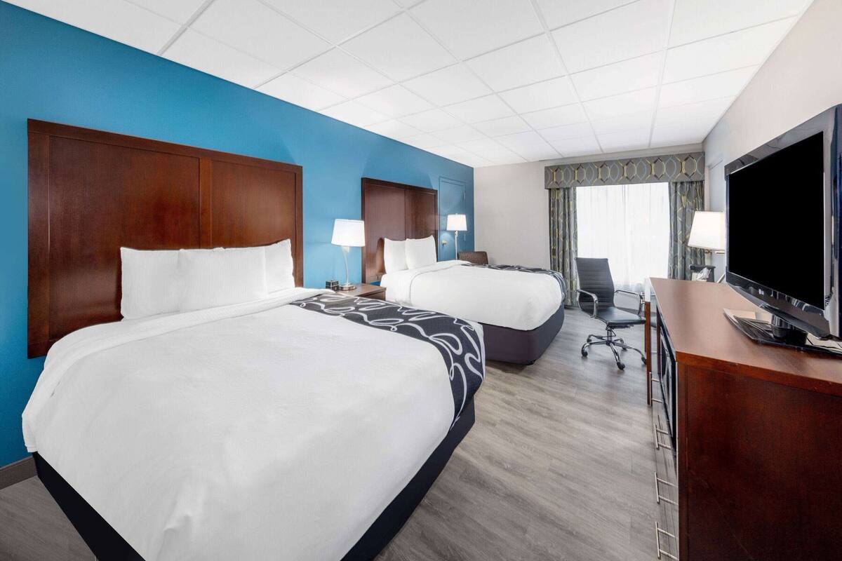 A hotel room with two beds, a desk with a TV, and a large window with curtains.