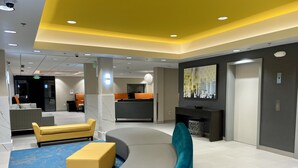 A modern lobby with a curved gray sofa, yellow ottoman, and teal armchair. There is a reception desk, a large TV screen, and a blue patterned carpet. - La Quinta Inn by Wyndham Lynnwood (Lynnwood)