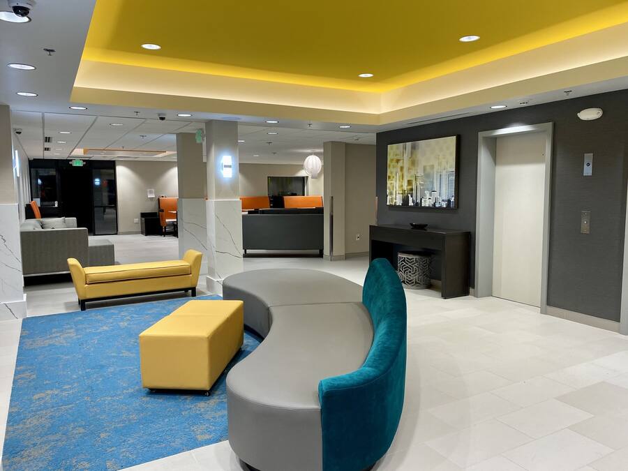 A modern lobby with a curved gray sofa, yellow ottoman, and teal armchair. There is a reception desk, a large TV screen, and a blue patterned carpet.