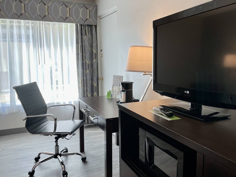 A hotel room with a desk, a chair, a television, a lamp, and a window with curtains.