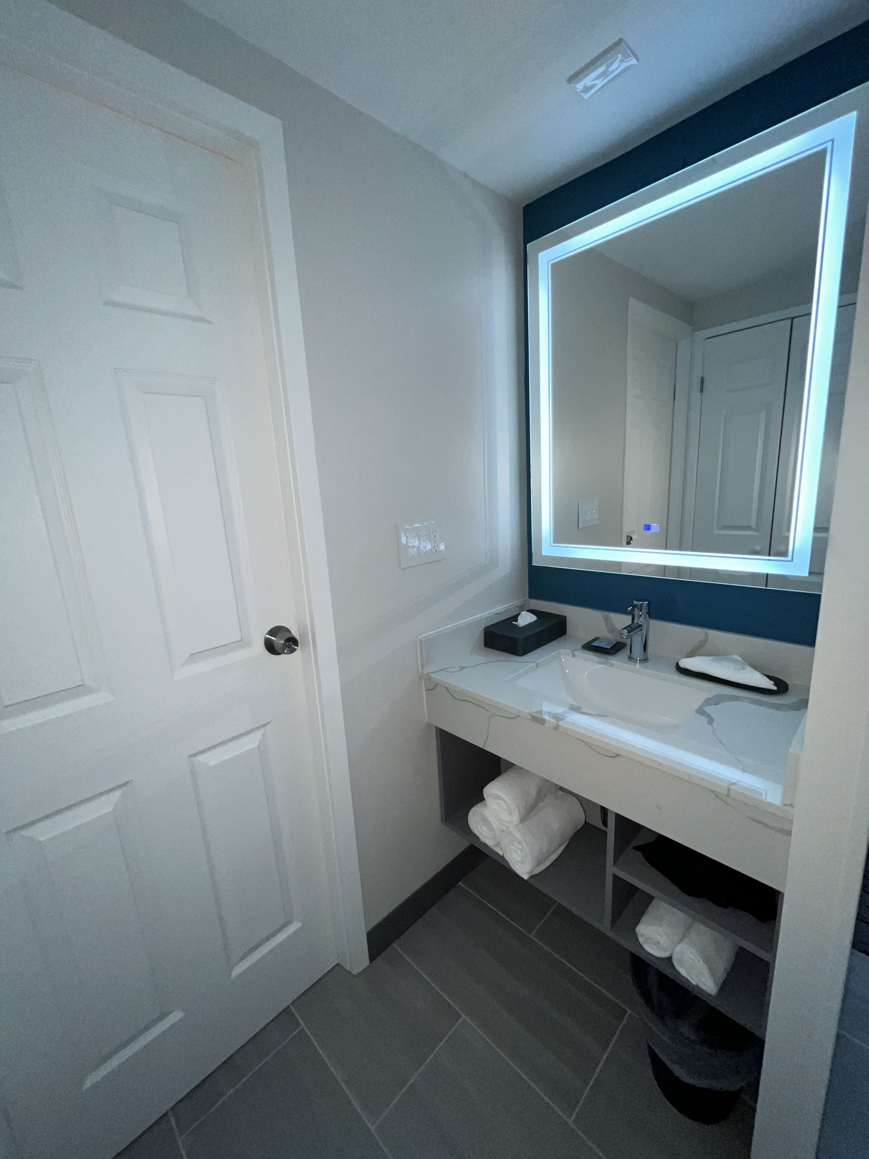 A bathroom with a white door, a sink with a mirror, and a towel rack.