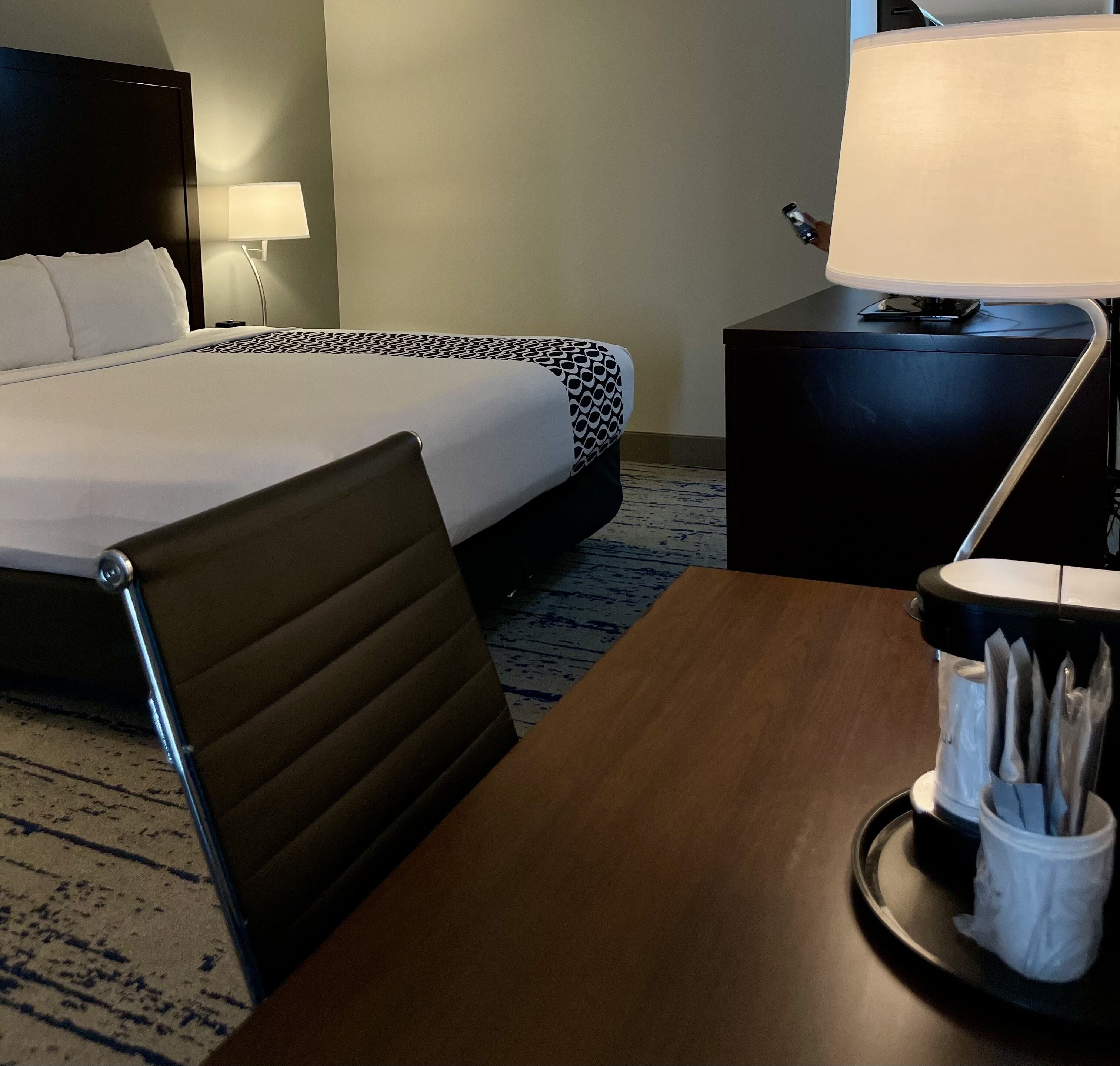 Habitación de hotel con cama, escritorio, silla, lámpara y cafetera.