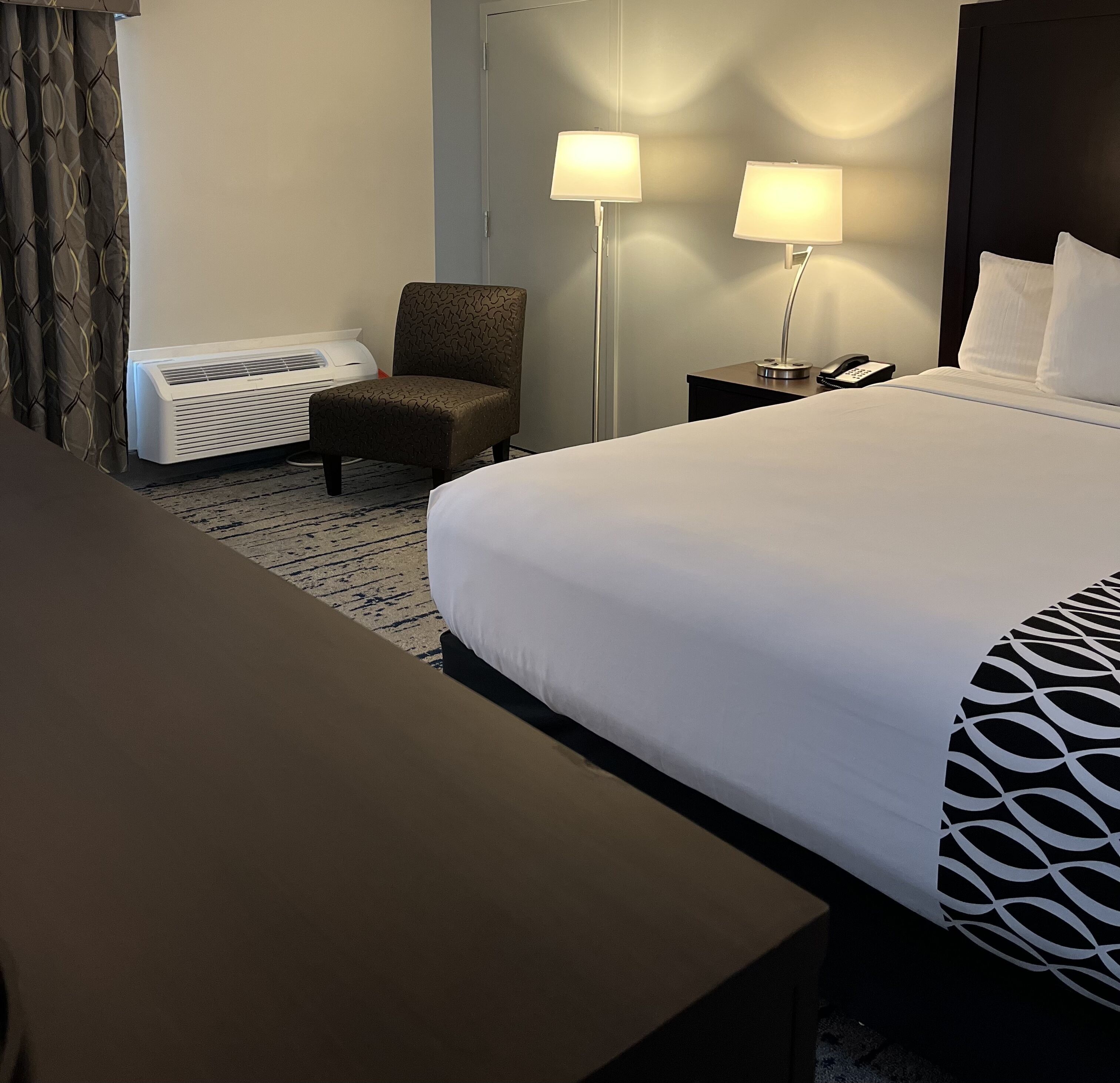 A hotel room with a large bed, a chair, a desk, two bedside lamps, a heater, and a telephone on a nightstand.