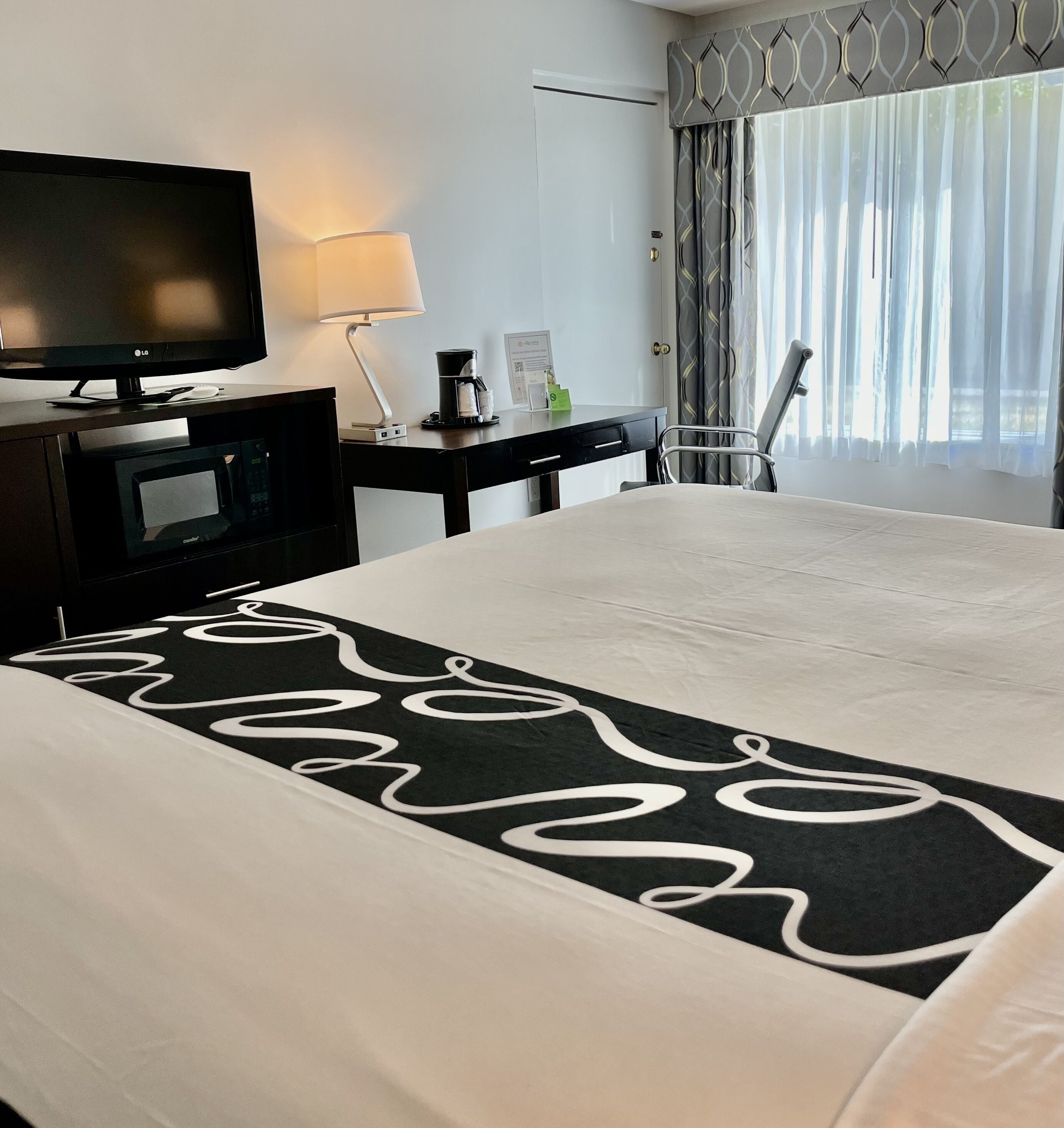 A hotel room with a bed, a TV, a desk with a lamp, a coffee maker, and a chair.