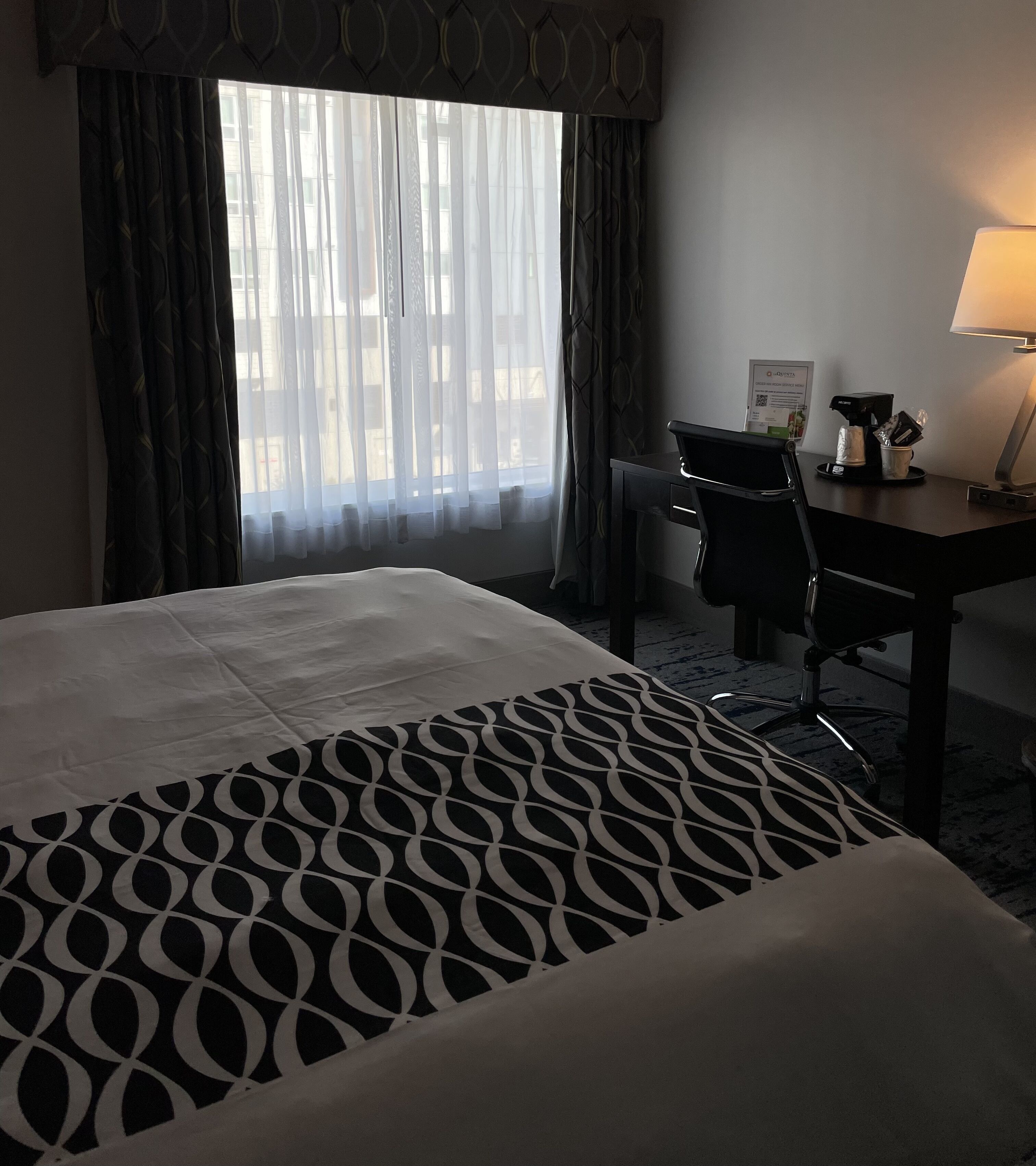 A hotel room with a large bed, a desk with a chair, a lamp, and a window with curtains.