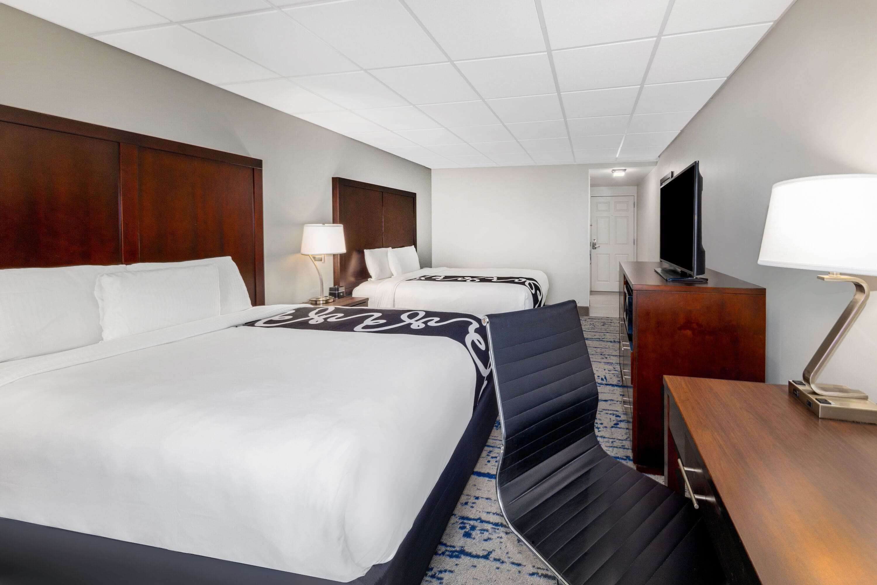 Habitación de hotel con una cama grande, una cama más pequeña, un escritorio con lámpara, televisión y armario.