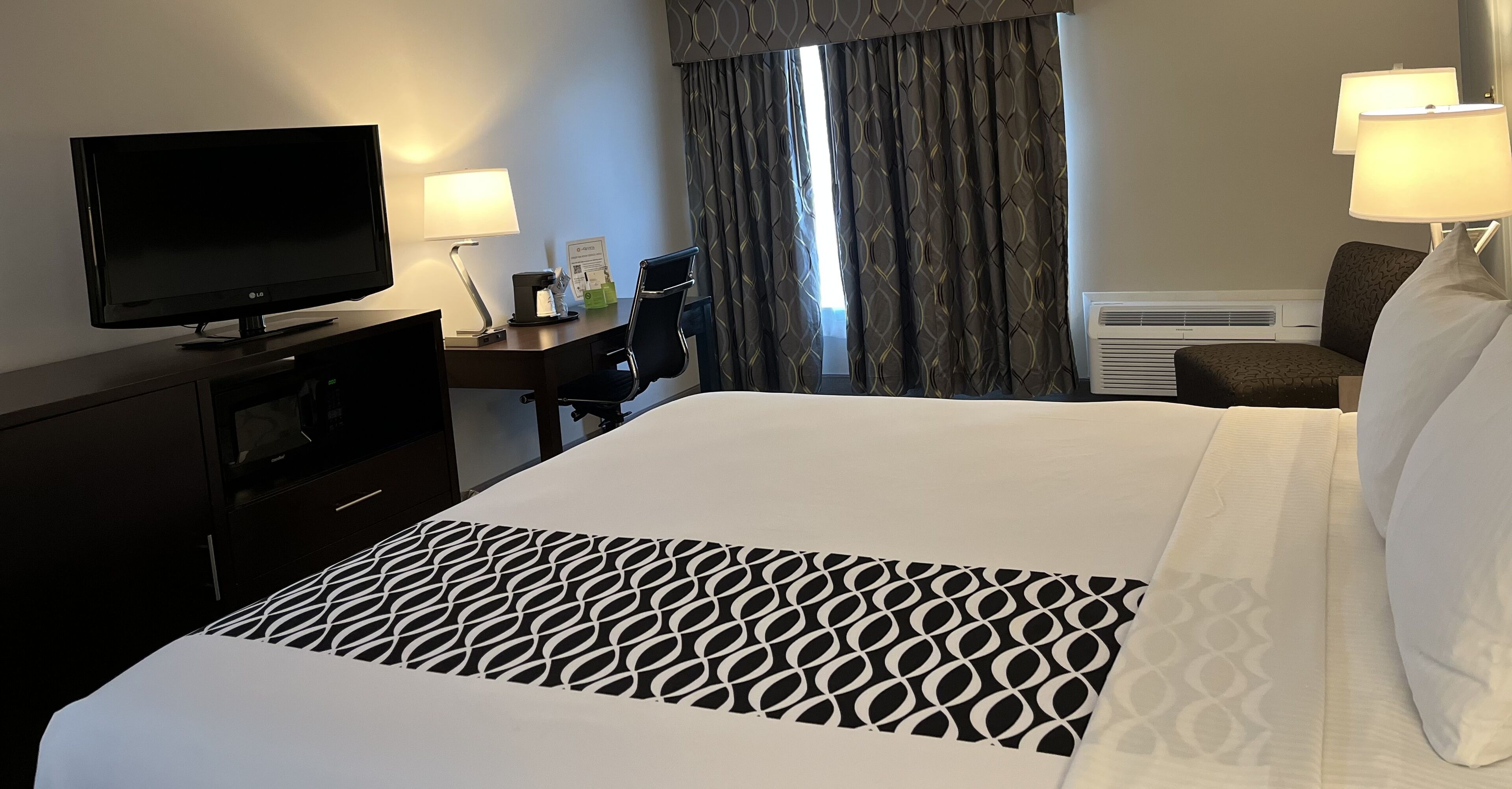 A hotel room with a bed, a desk with a computer, a television, and a window with curtains.