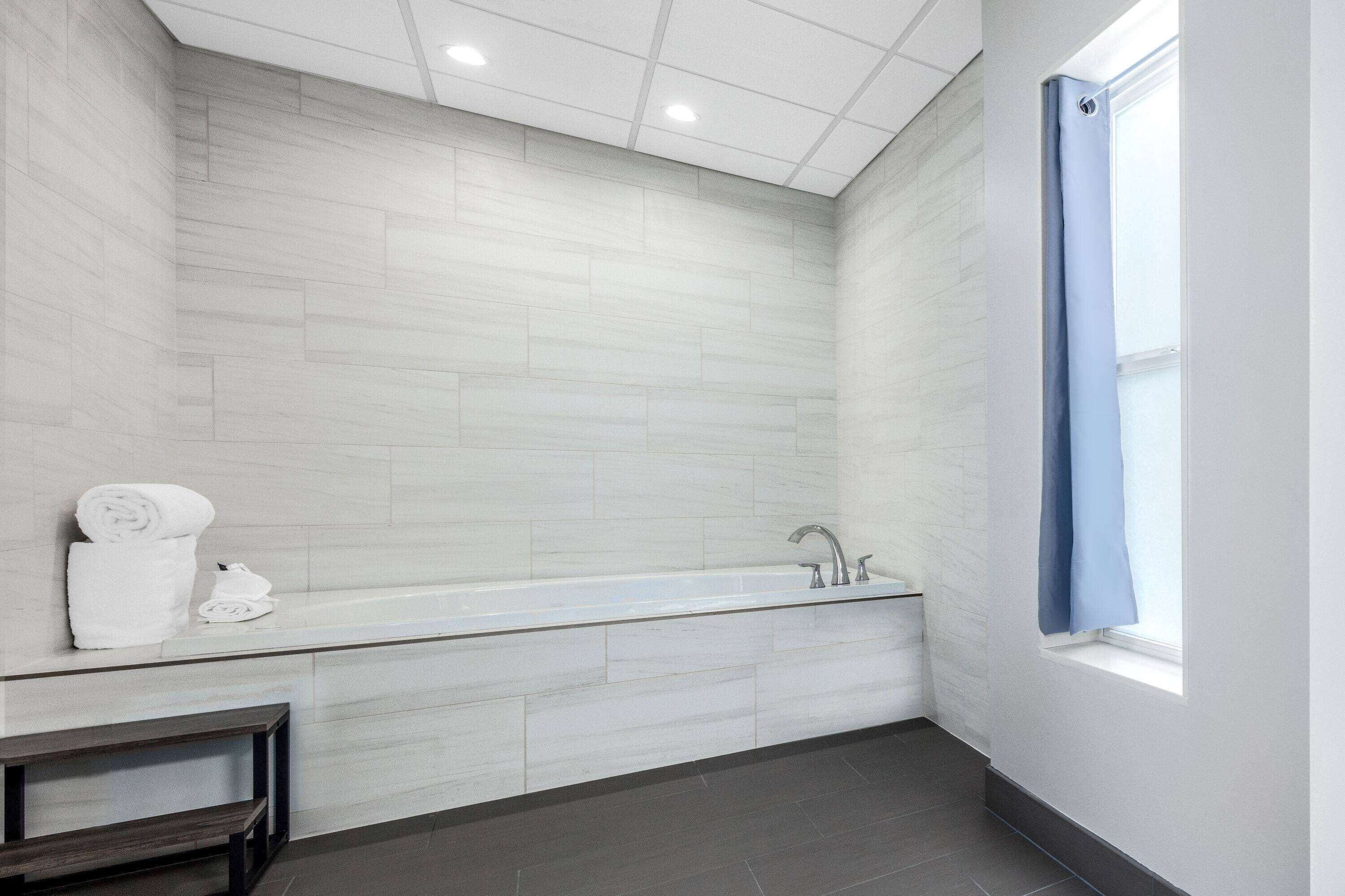 Un baño moderno con una bañera grande, un banco y un ventanal con cortina azul.