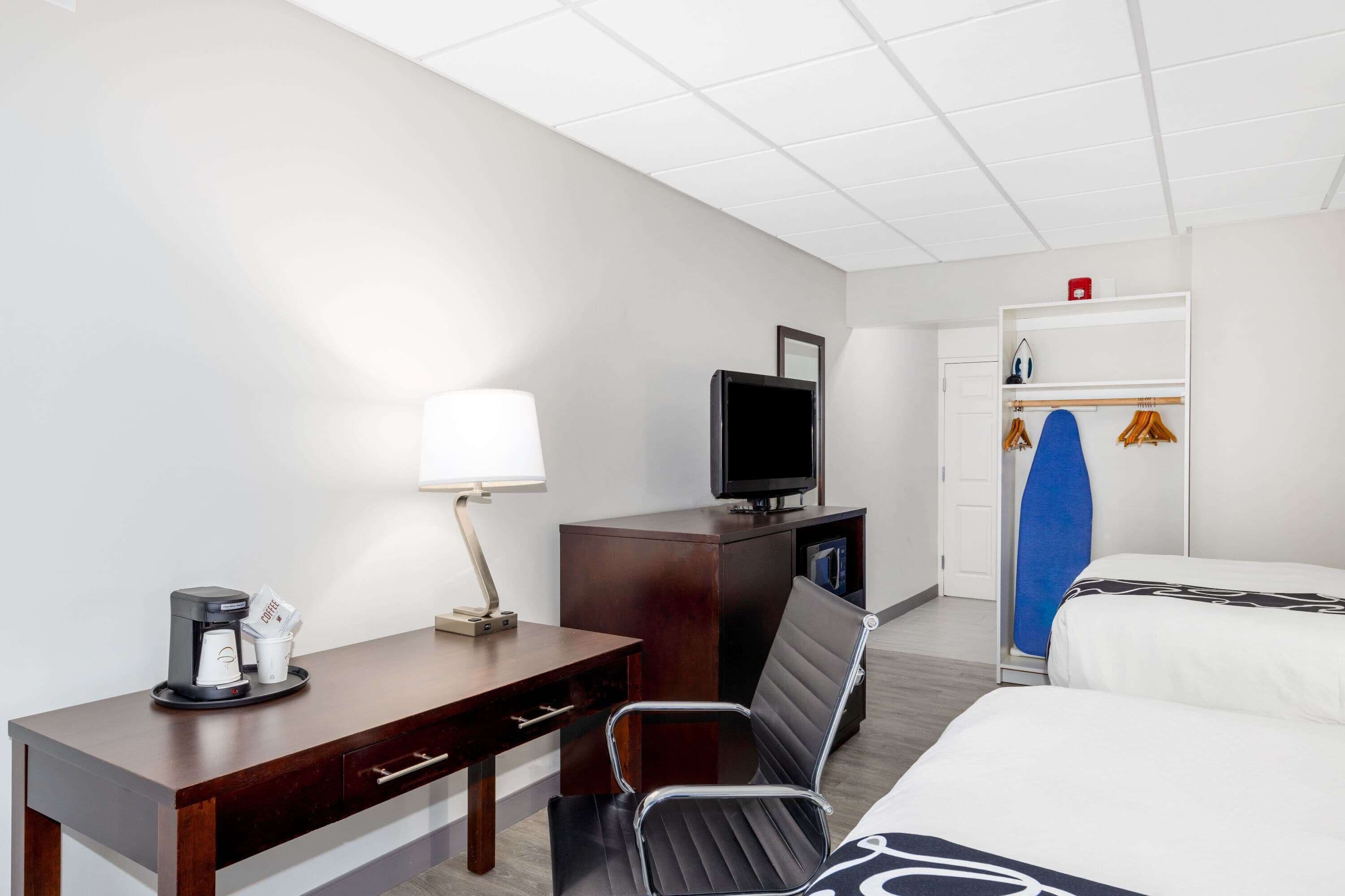 A hotel room with a bed, a desk with a lamp, a television, a coffee maker, and a wardrobe with a surfboard.
