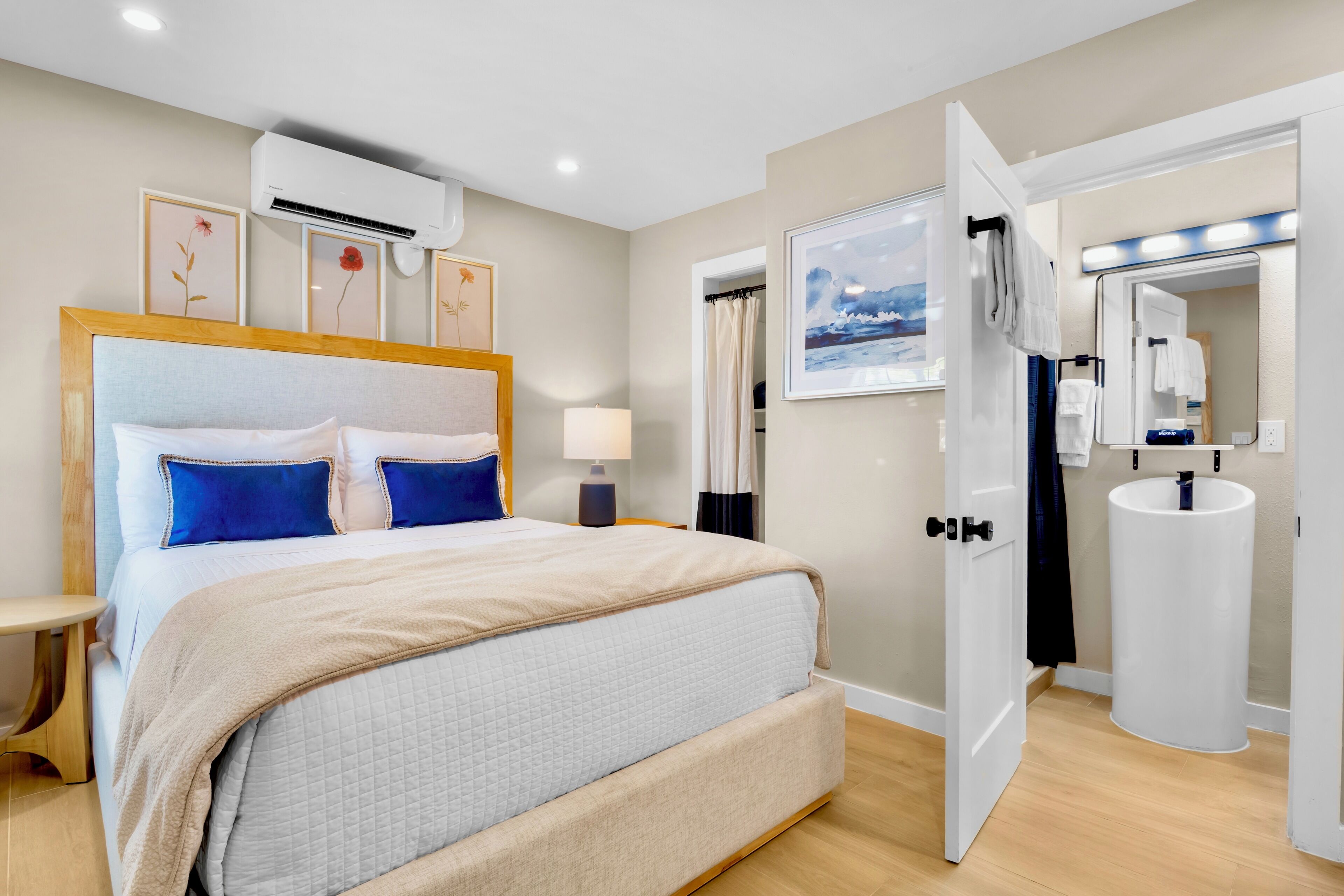 A hotel room with a bed, bedside table, lamp, air conditioning unit, framed pictures, a bathroom with a sink, mirror, and towel rack.