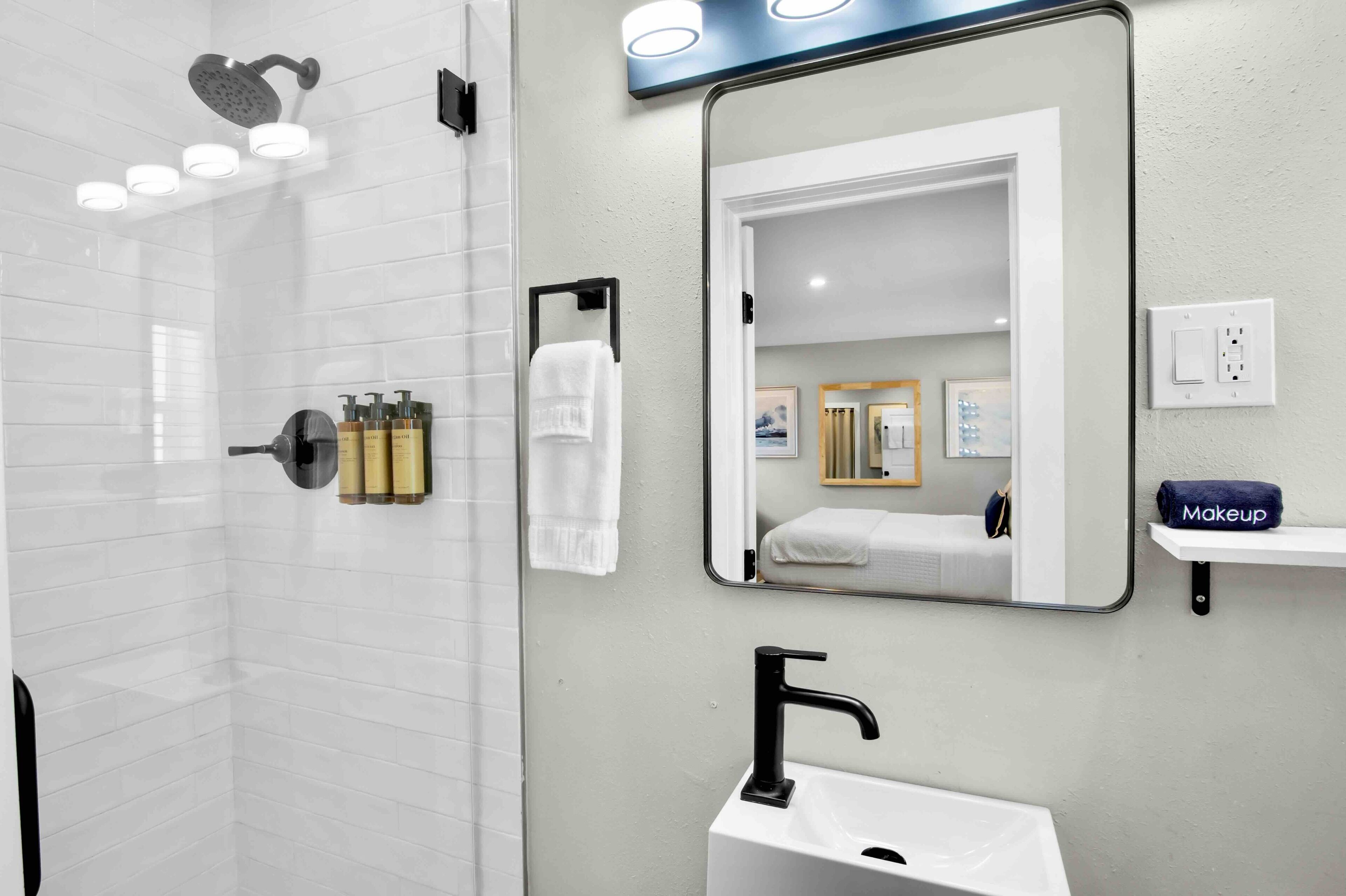 Une salle de bain moderne avec une douche à parois de verre, un lavabo blanc, un grand miroir et un meuble-lavabo doté d’une étagère pour les produits de maquillage.