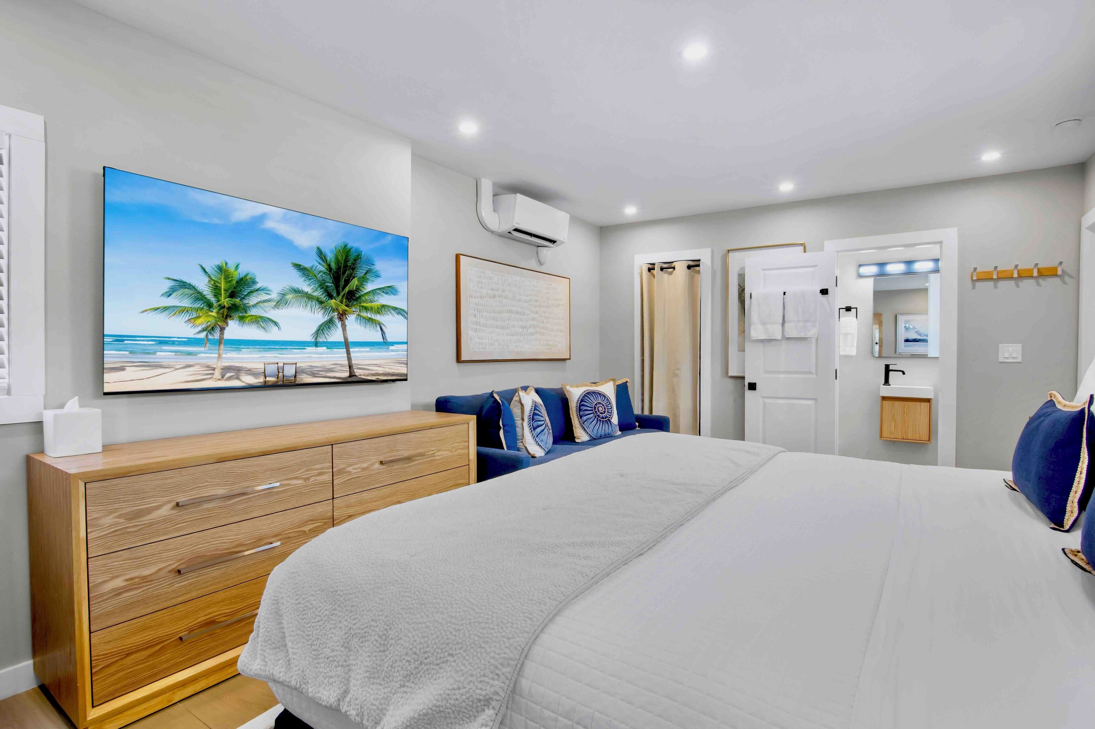 A bedroom with a large bed, a wooden dresser, a painting of a beach, and a bathroom with a shower curtain.