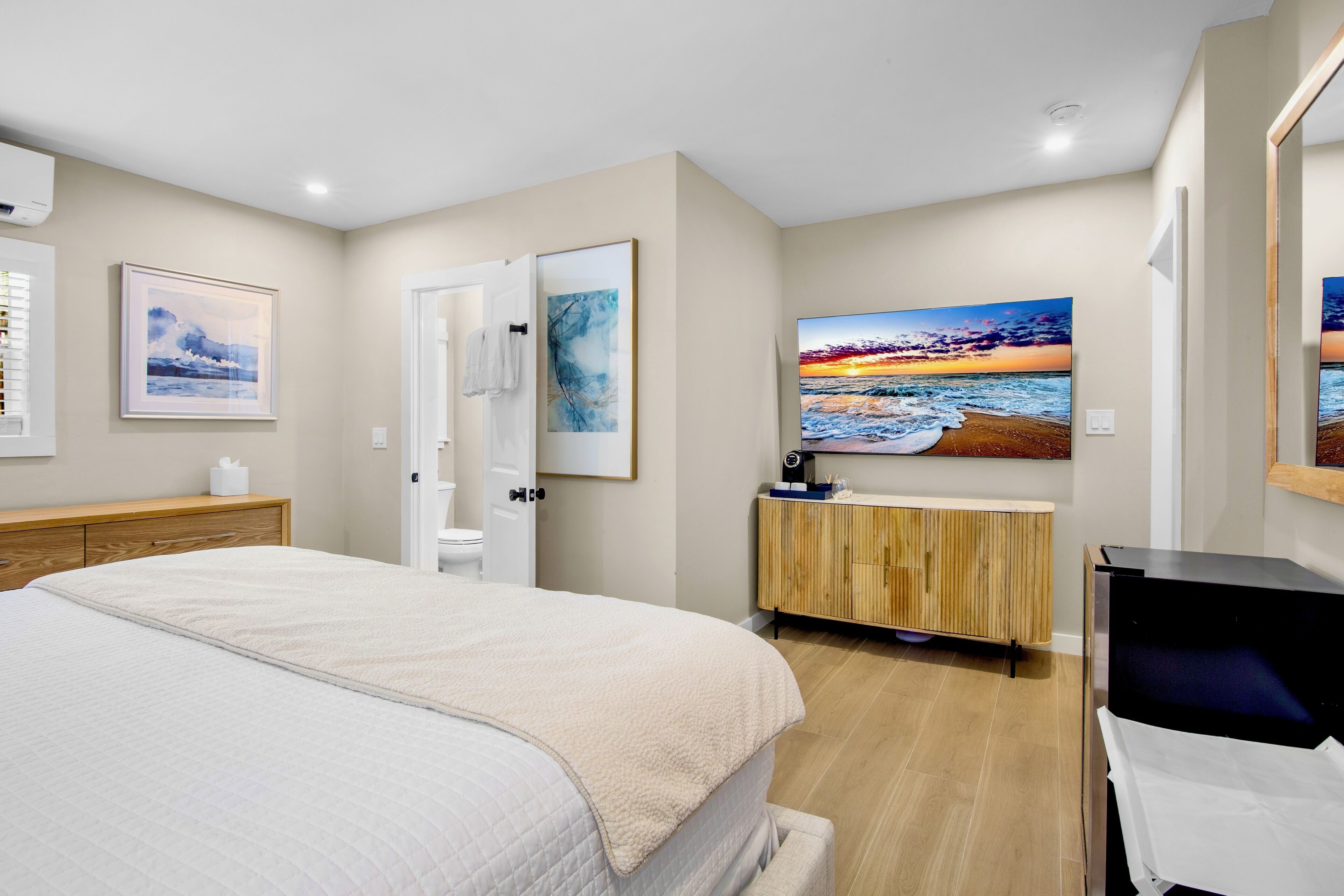 A bedroom with a bed, a wooden dresser, a vanity, and a large painting of a sunset over the ocean.