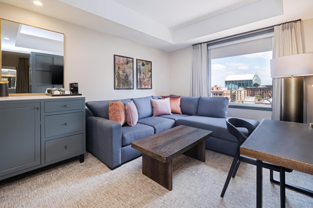 A modern living room with a grey sofa, a wooden coffee table, a dining table, and a grey cabinet. There are two framed artworks on the wall, a large mirror, and a window with a view of buildings.