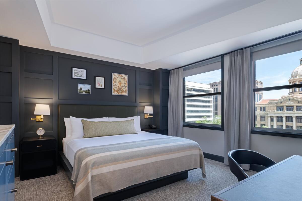 A hotel room with a large bed, two bedside tables with lamps, a desk, and a view of a building through the window.