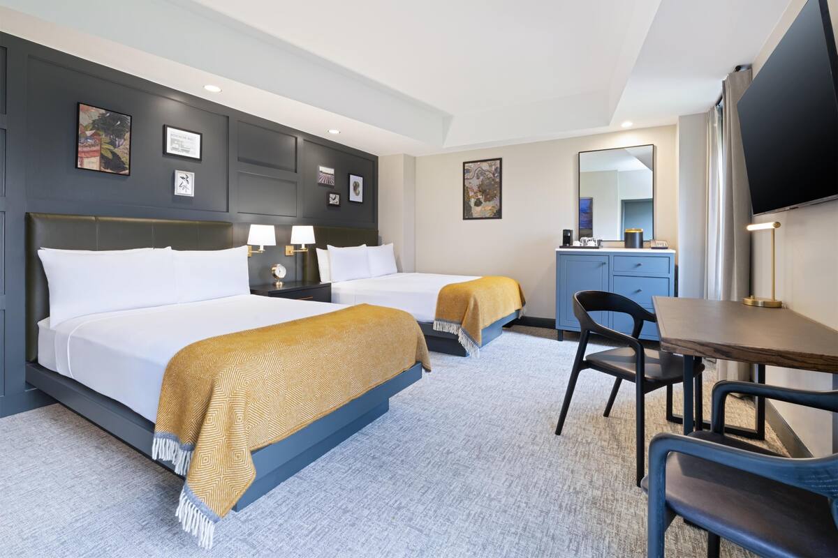 A hotel room with two beds, a desk, a chair, a TV, and a minibar.