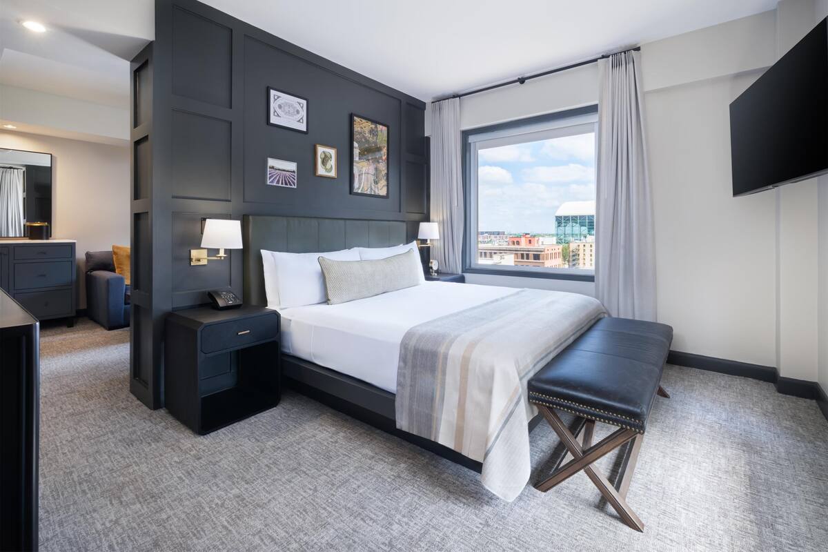 A modern hotel room with a large bed, bedside tables, a bench, a TV, and a city view.