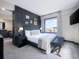 Habitación de hotel moderna con una cama grande, mesitas de noche, un banco, televisión y vistas a la ciudad.