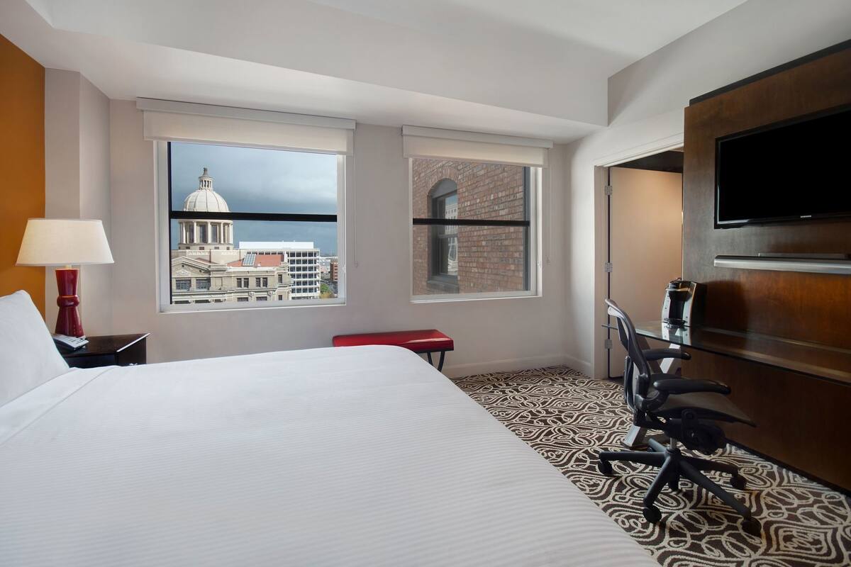 A hotel room with a bed, a desk with a chair, a TV, and two windows offering city views.