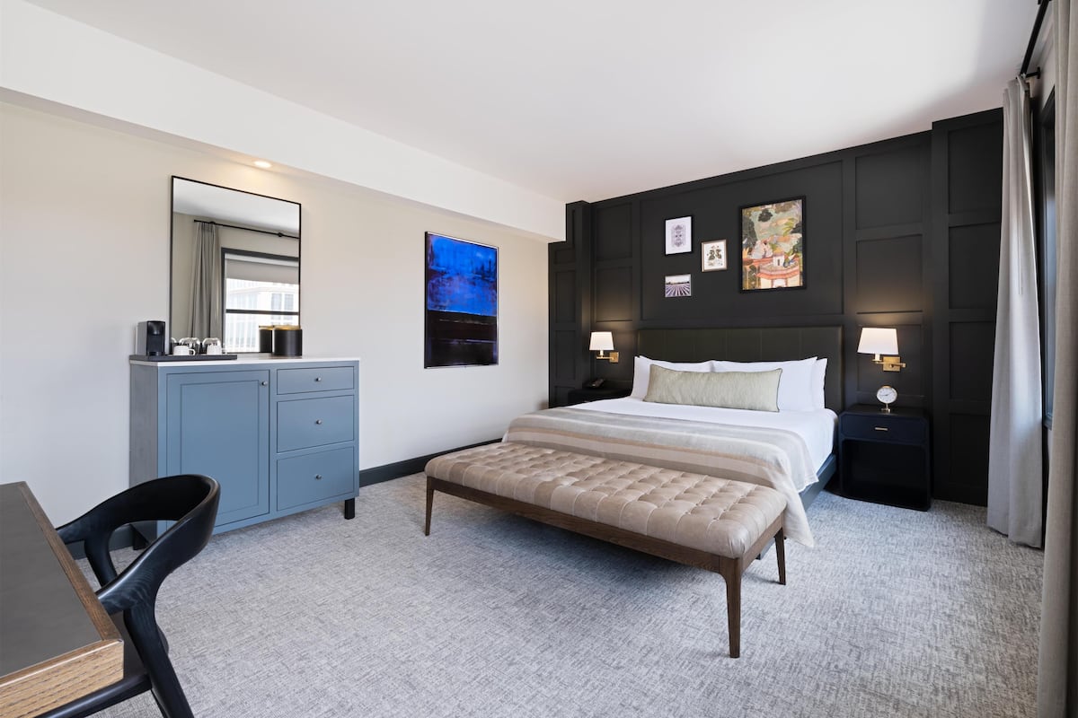 A hotel room with a bed, a desk, a chair, a dresser, and a wall with artwork.