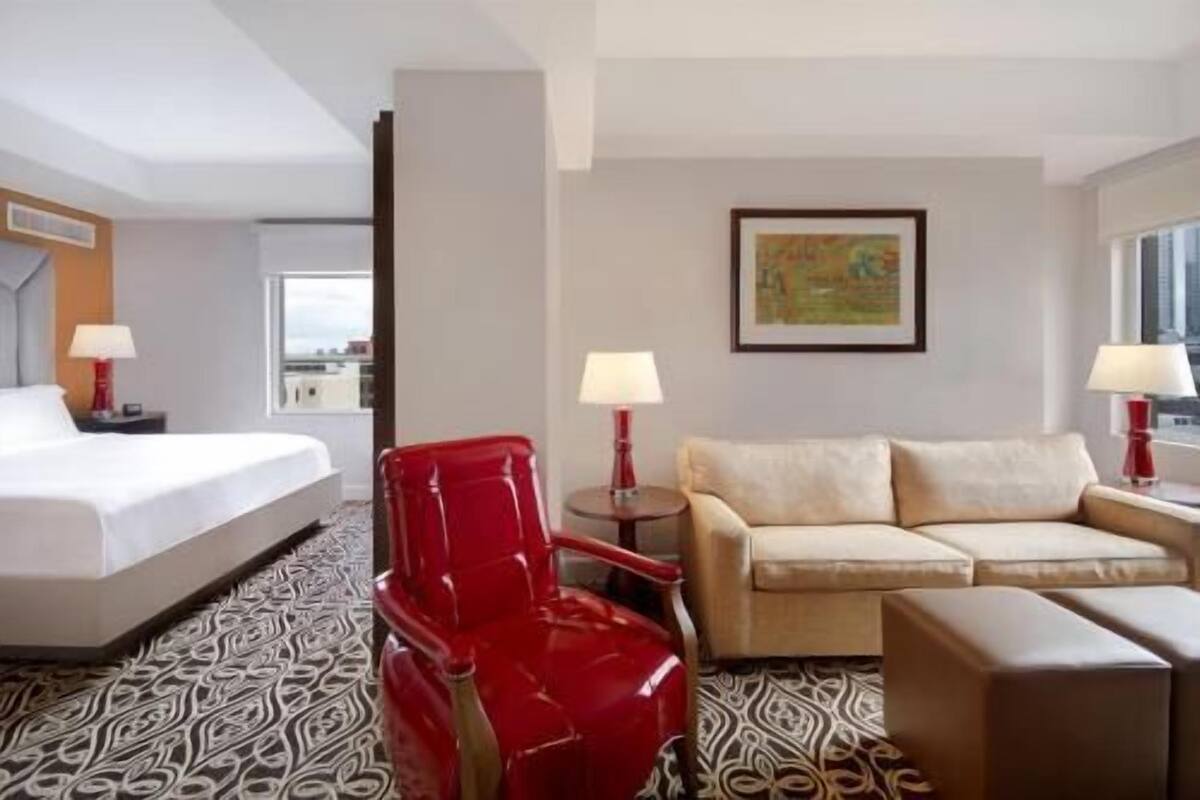 A hotel room with a bed, a red chair, a sofa, two lamps, and a painting on the wall.