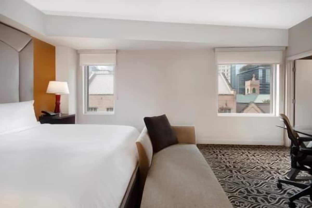 A hotel room with a bed, a desk with a lamp, a chair, a patterned carpet, and a window with a city view.