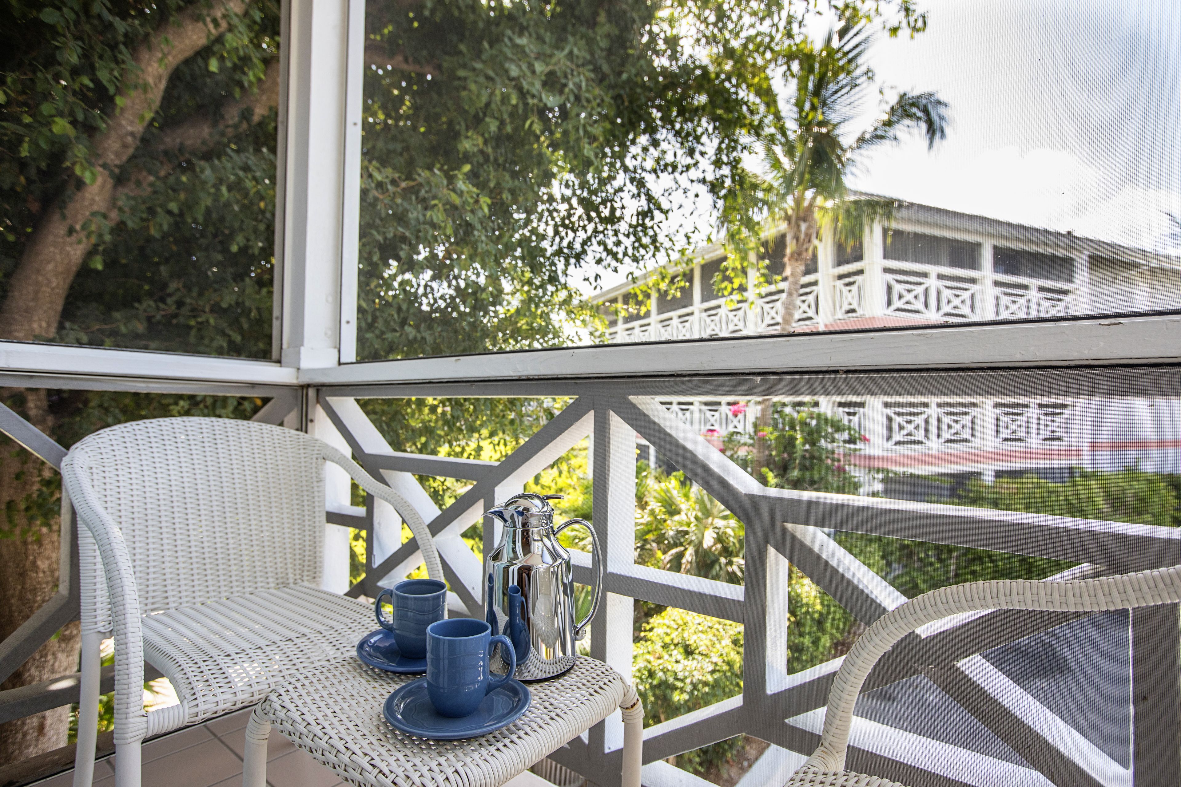 Una silla de mimbre blanca con una mesa, una jarra y dos tazas azules en un balcón con vistas a la vegetación y un edificio blanco.