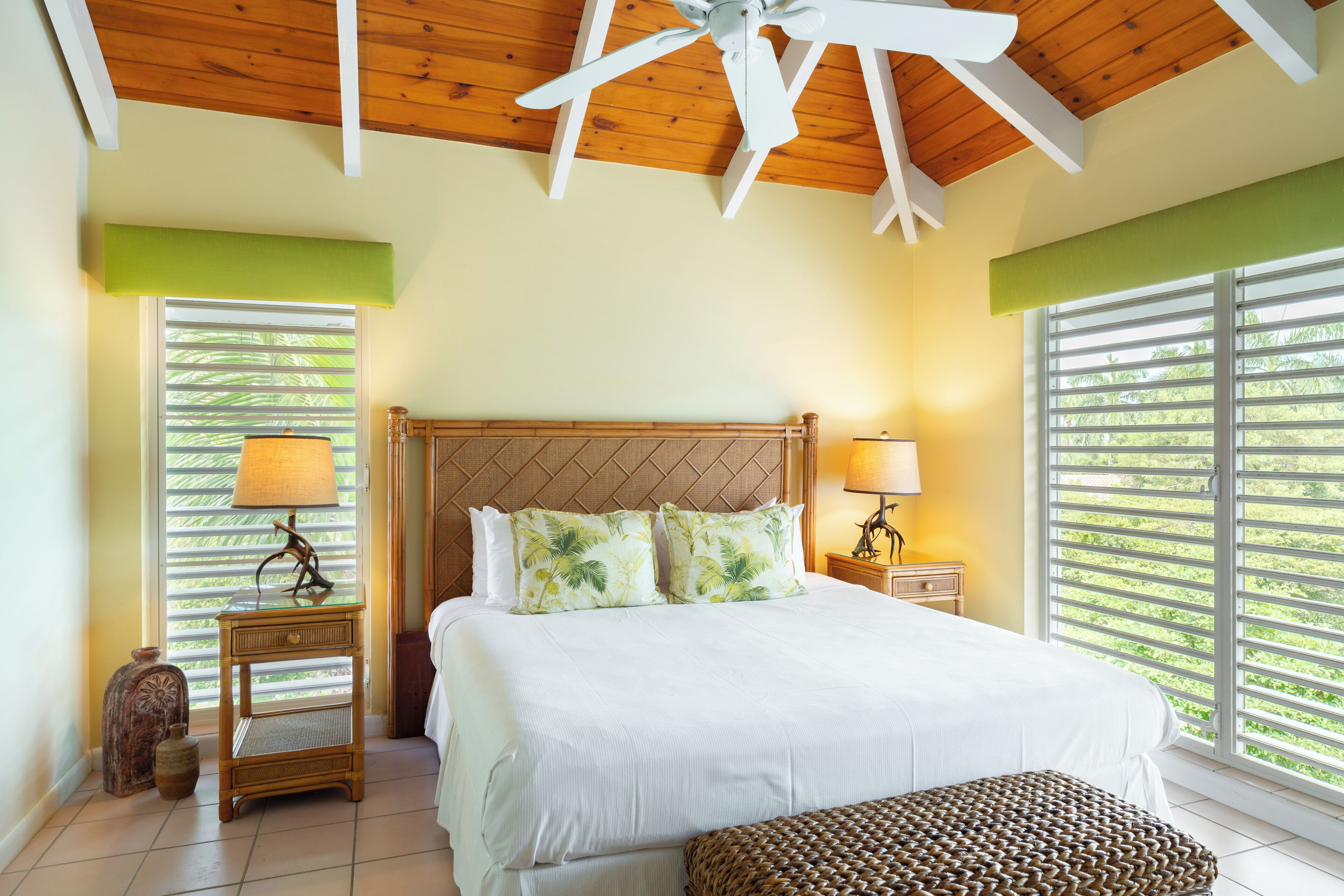 A bedroom with a large bed, wooden ceiling, two nightstands with lamps, a wicker bench, and large windows with blinds.