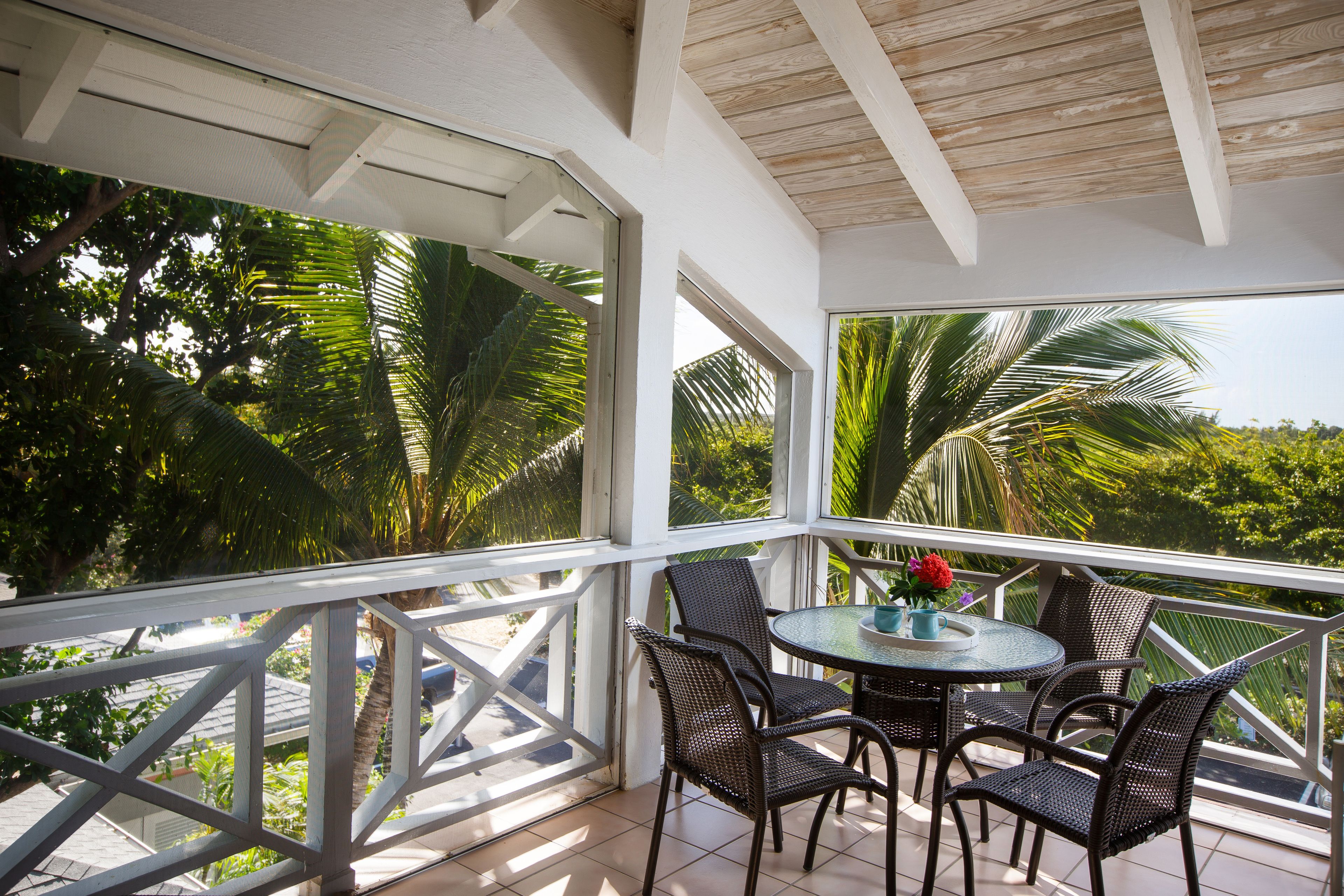 Un espace extérieur abrité, doté d’une table ronde et de quatre chaises, et entouré de grandes fenêtres offrant une vue sur les palmiers et la végétation.