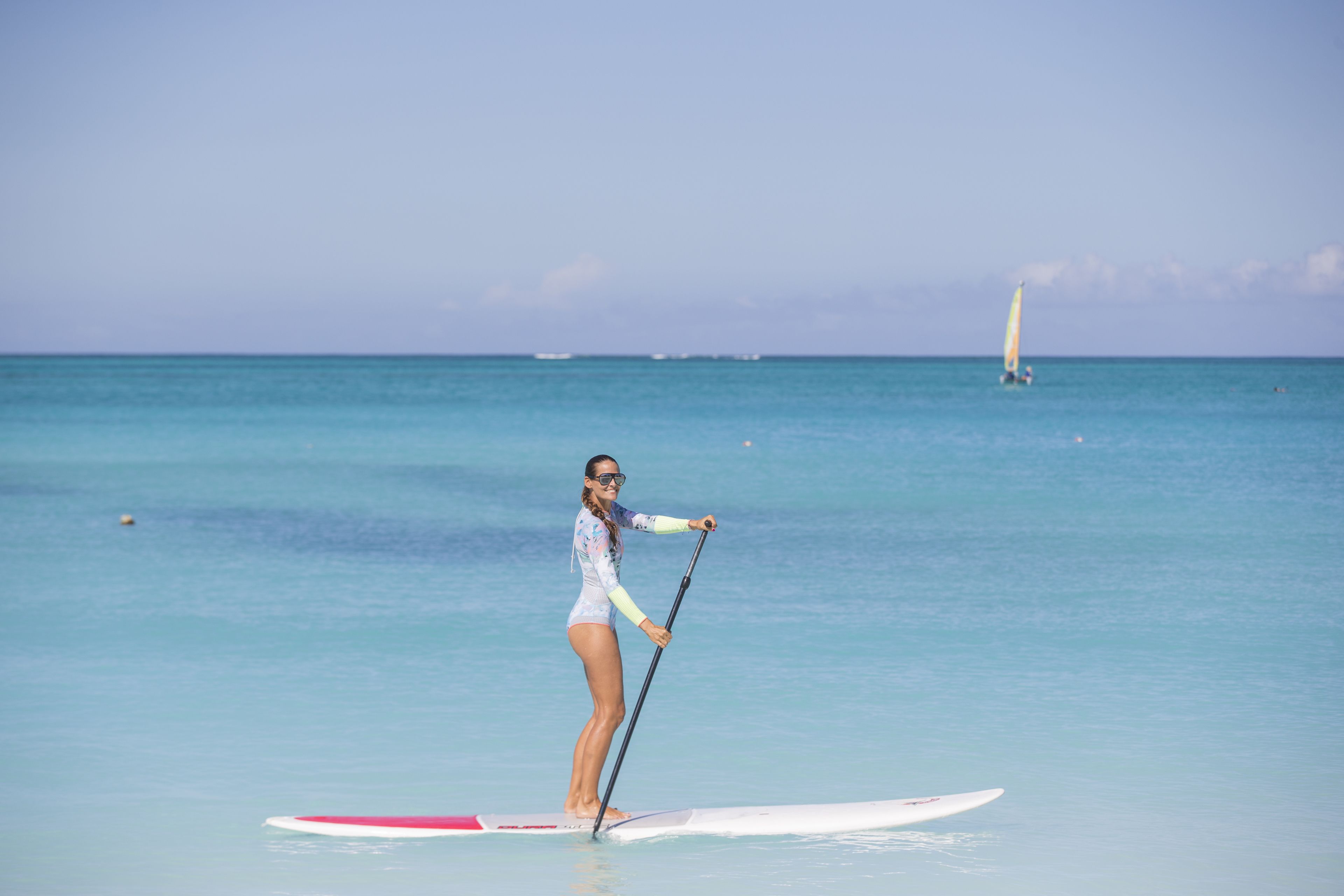A person paddleboarding on a calm sea with a sailboat in the background.