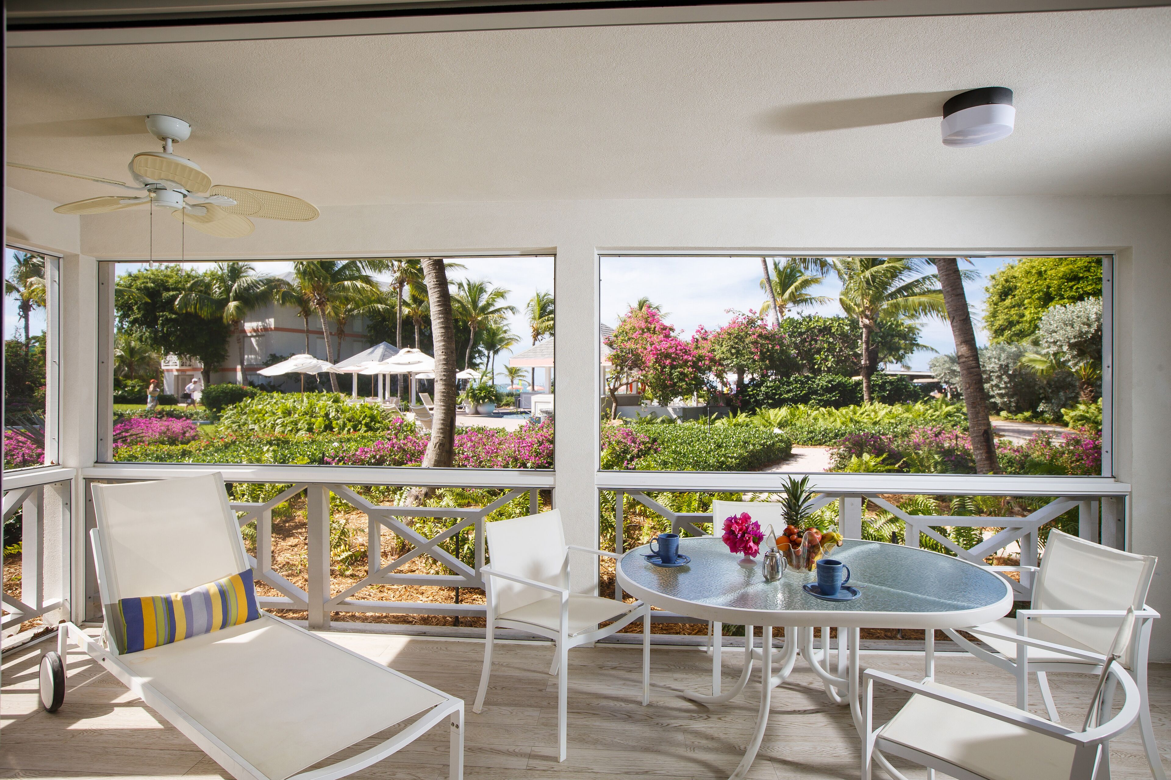 A sunlit outdoor patio with white furniture, a round table, and a view of a lush garden and palm trees.