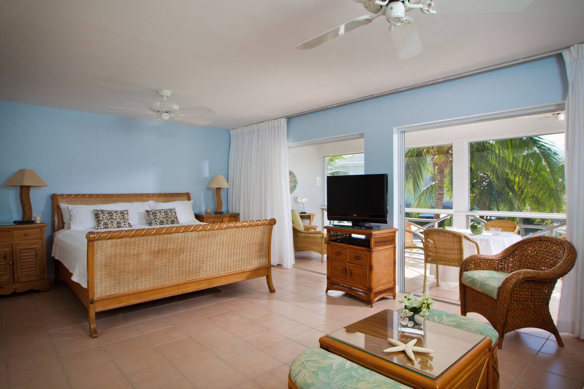 Quarto com cama, televisão, móveis de vime e varanda com vista para palmeiras.