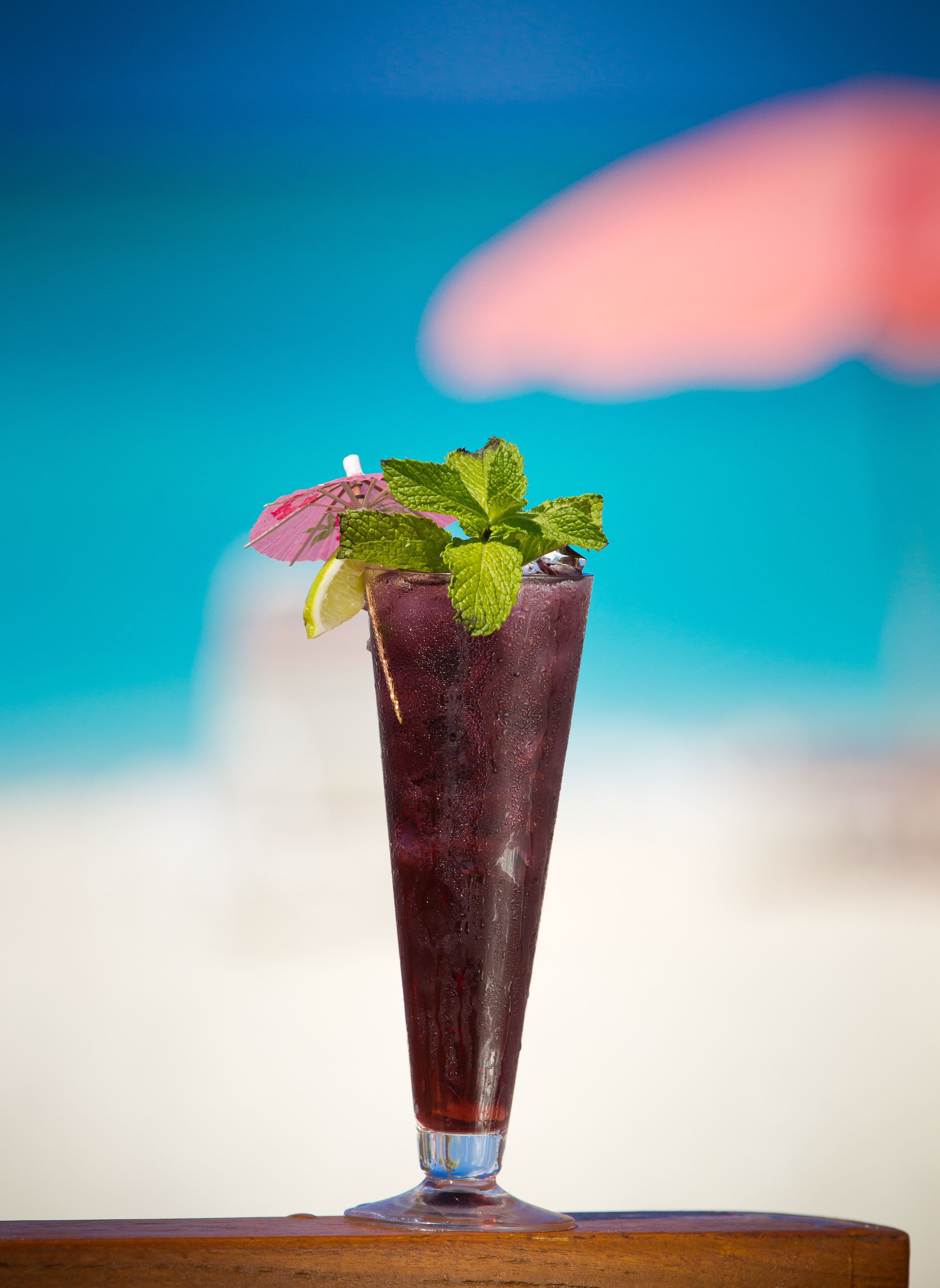 A tall glass with a dark-colored beverage, garnished with mint leaves and a slice of lime, on a wooden surface against a beach backdrop.