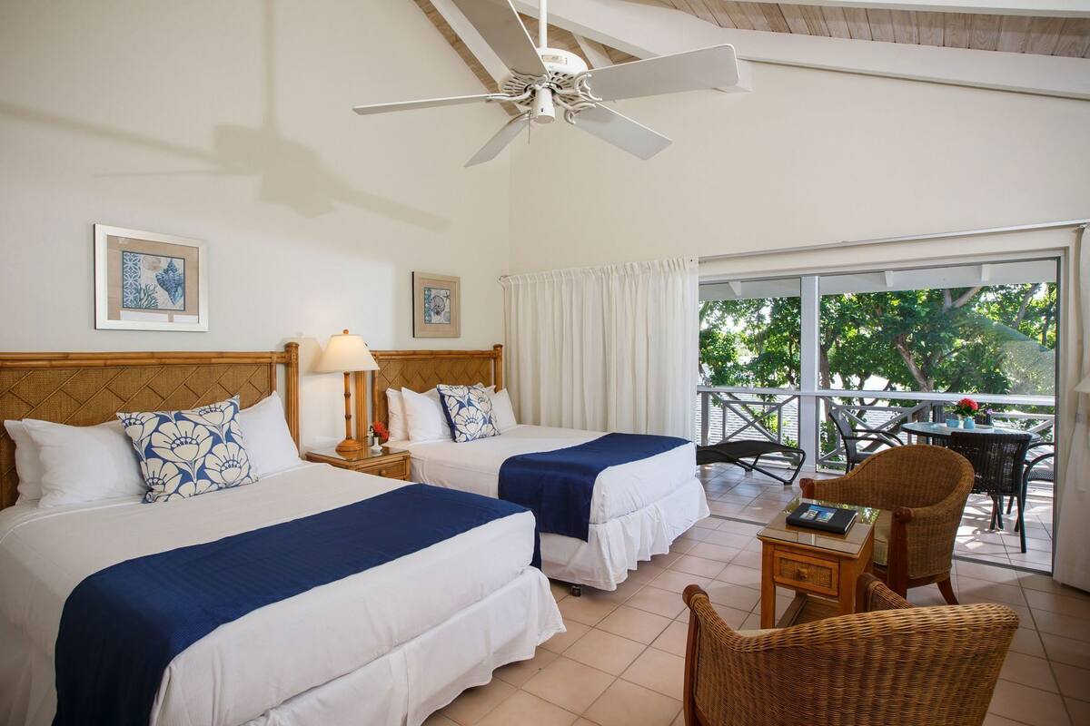 Quarto de hotel com duas camas, um ventilador de teto, uma varanda com mobiliário exterior e vista para vegetação.