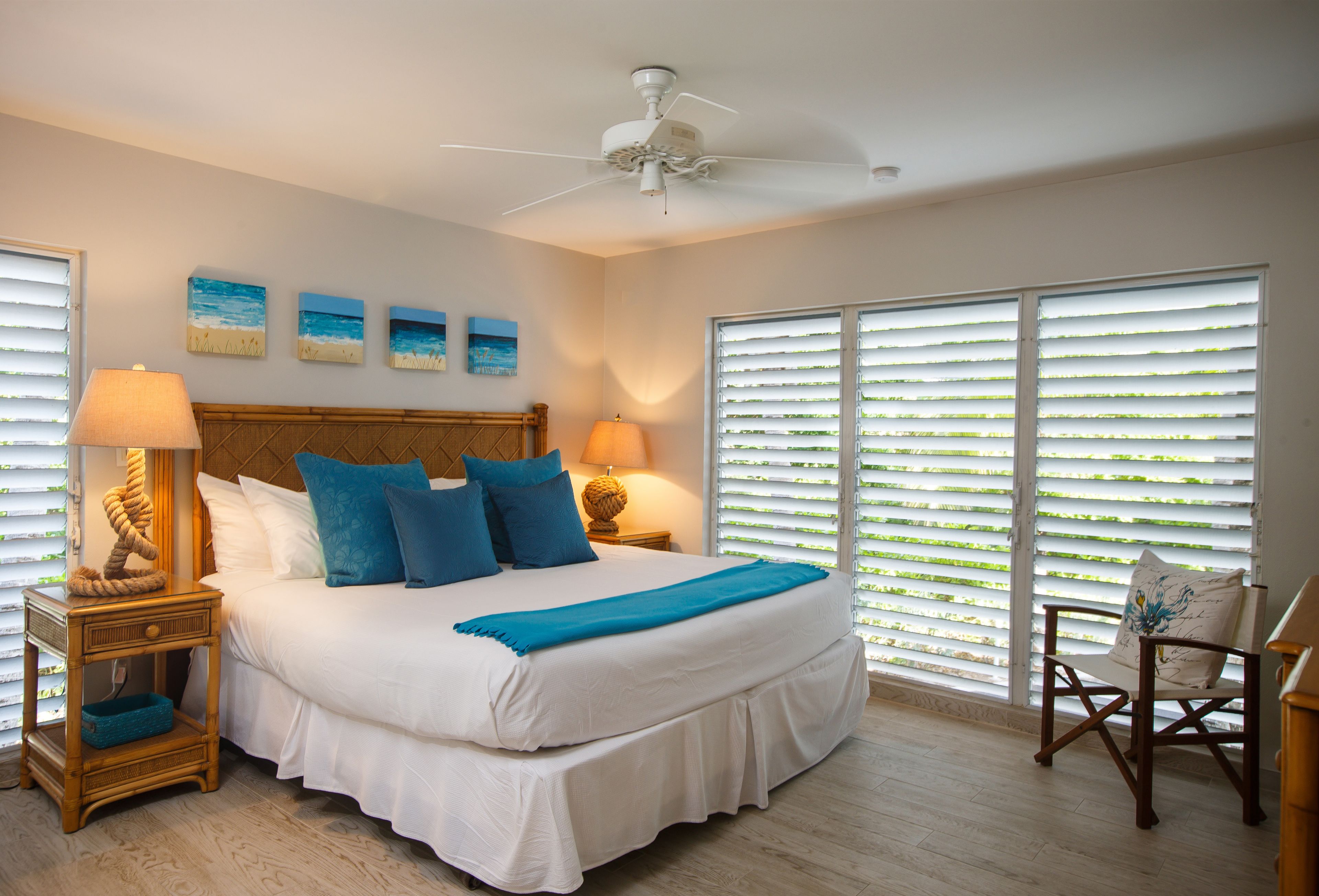 A bedroom with a large bed, two bedside tables with lamps, a ceiling fan, and a view through large windows with blinds.
