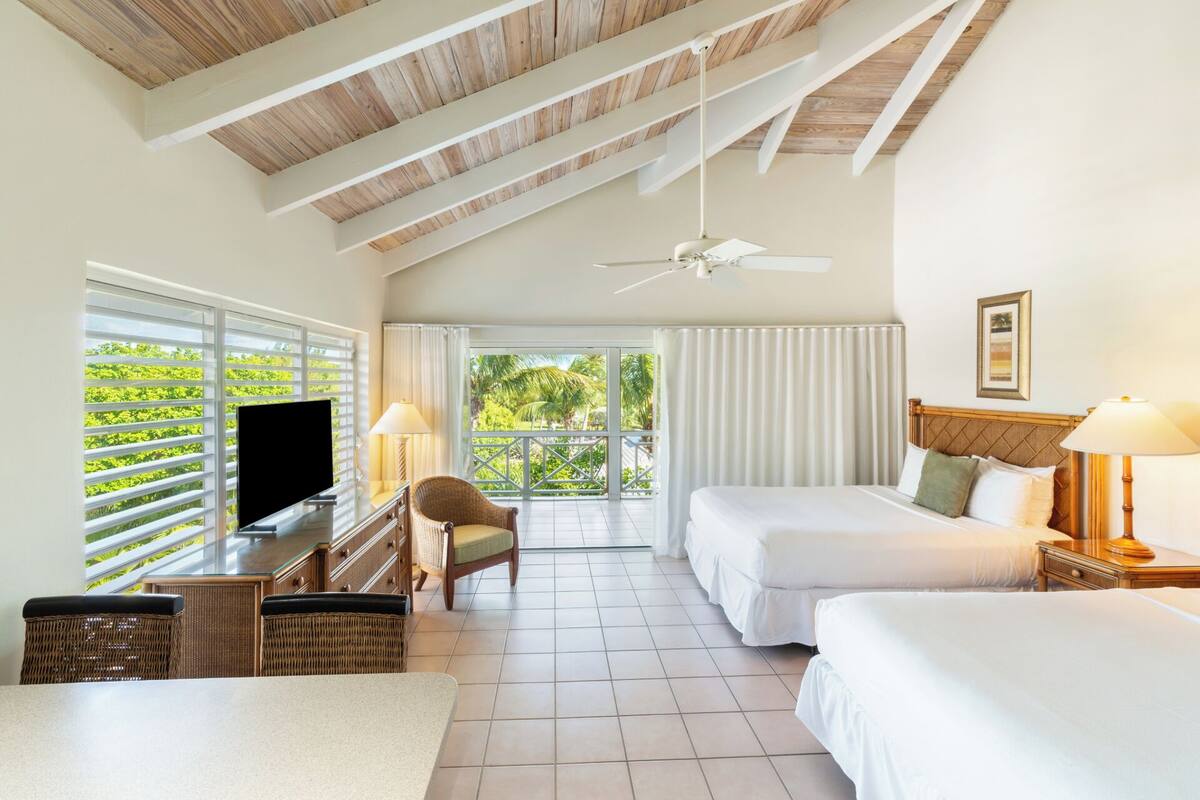 Quarto de hotel com uma grande janela, ventoinha no teto, duas camas, televisão e área de refeições com mobiliário de vime.
